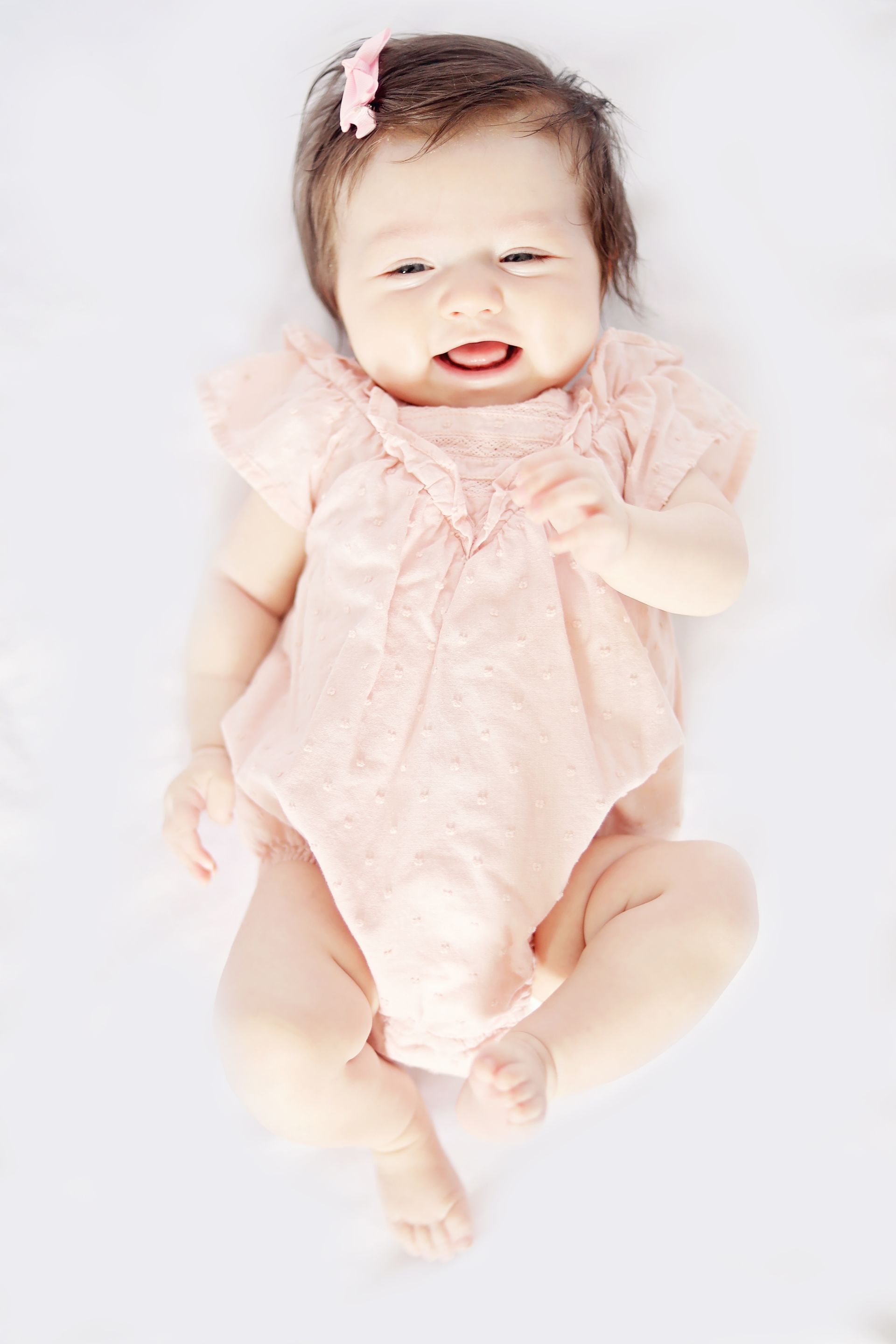 Baby in a light pink outfit lying on a white blanket, smiling with a bow in her hair