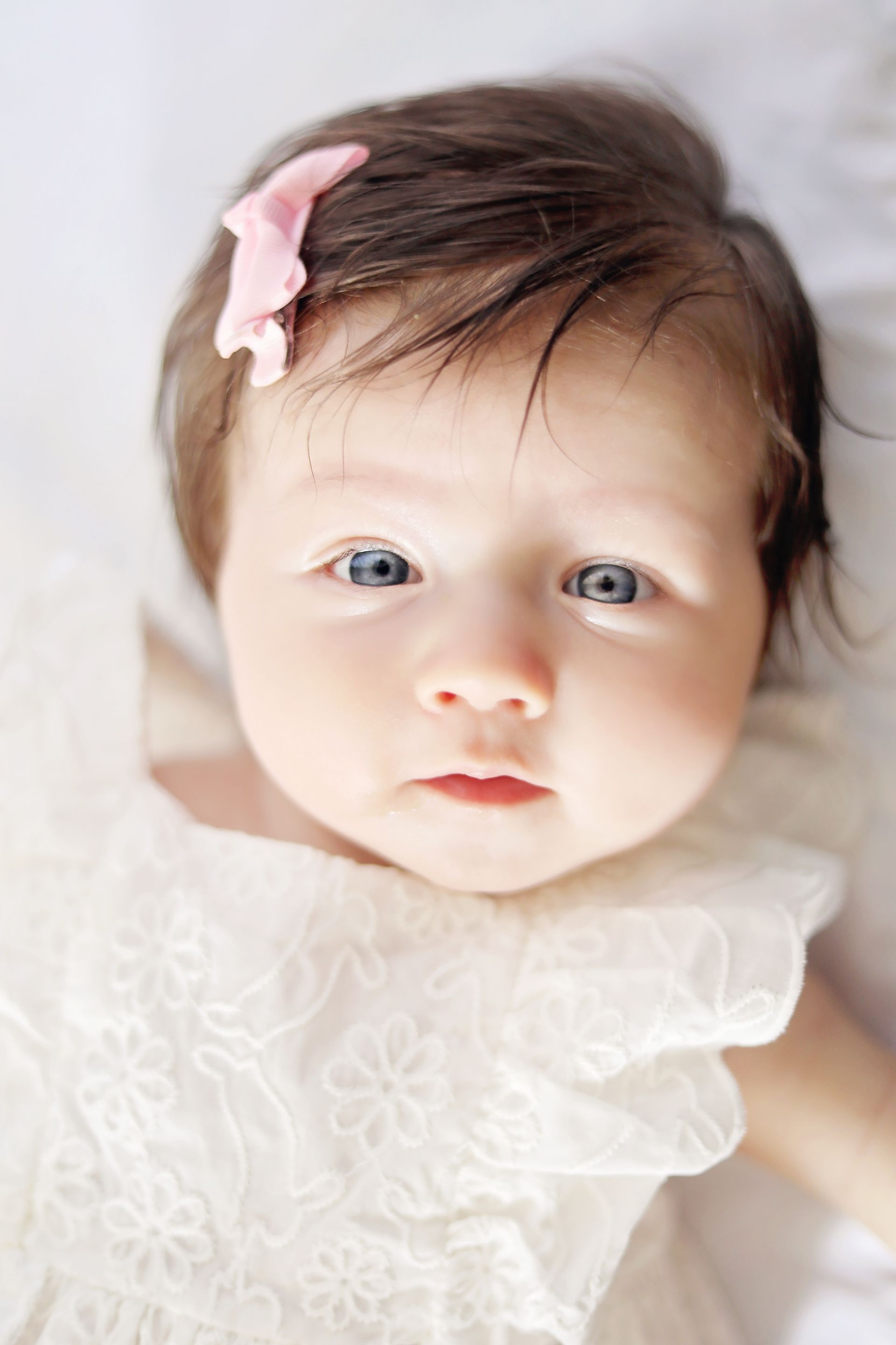 Close-up of a baby with a pink bow, wearing a white lace outfit and looking at the camera.