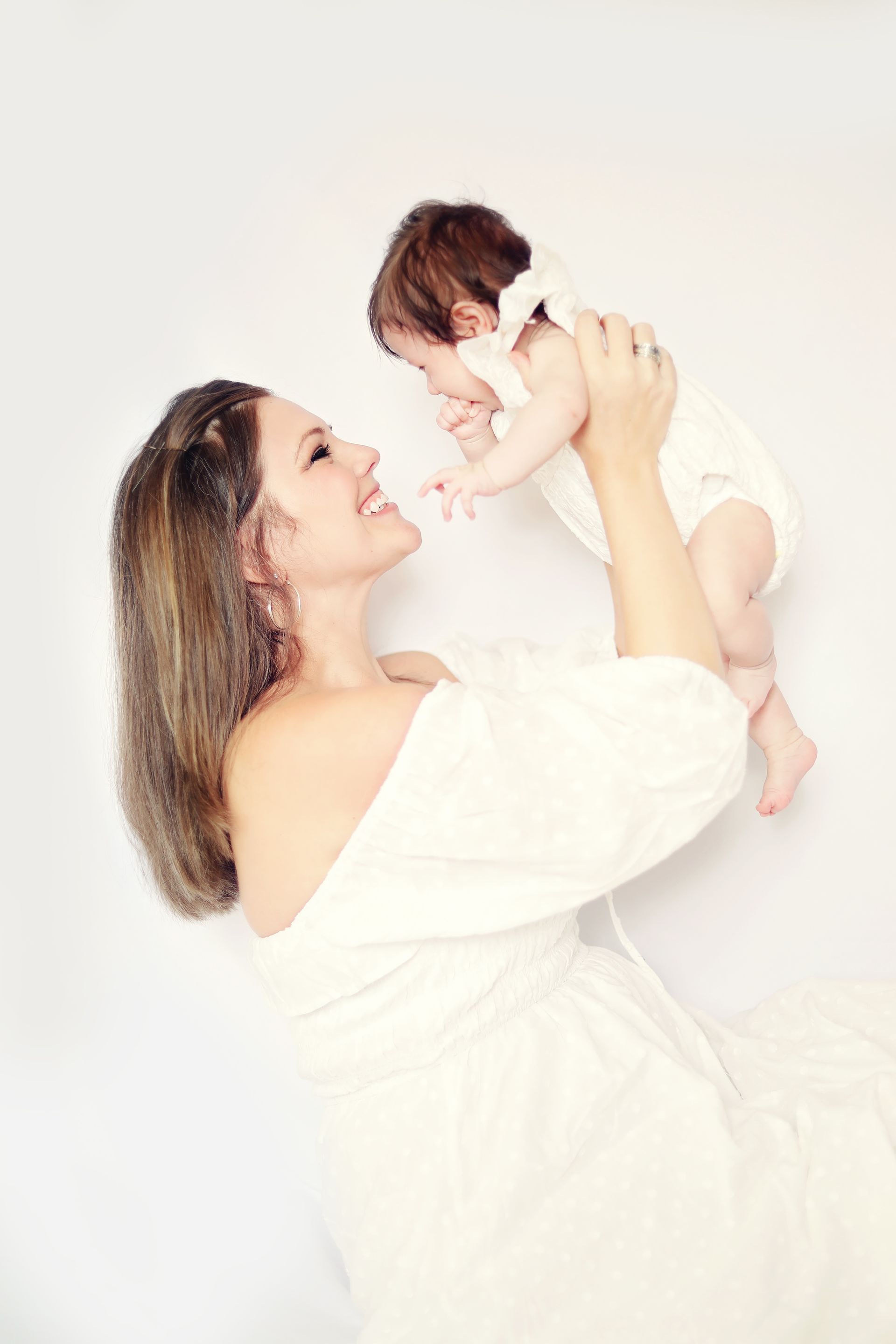 Woman in a white dress holding a baby in a bright studio, both looking at each other lovingly