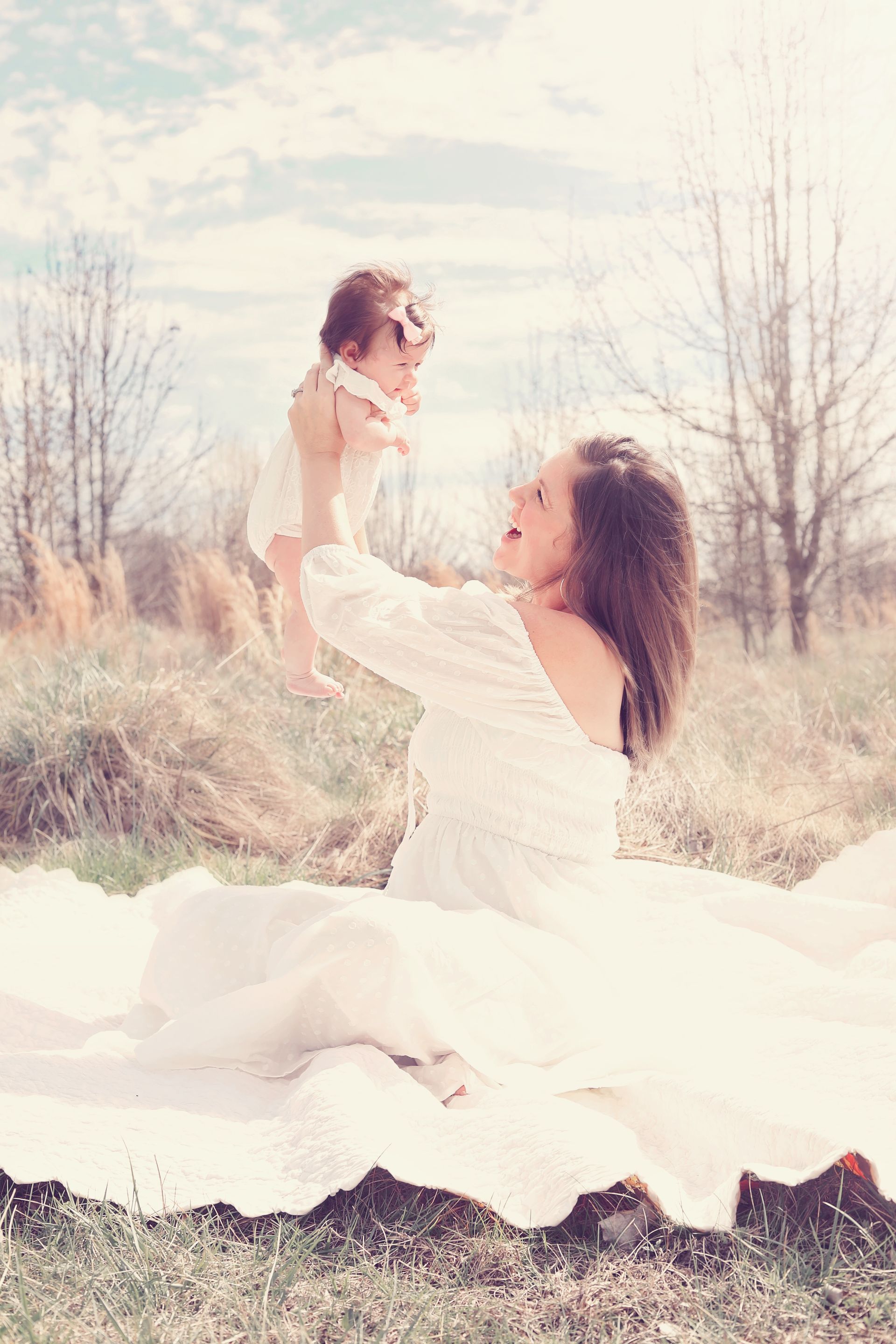 Woman lifting a toddler outdoors in a soft, sunlit field with bare trees