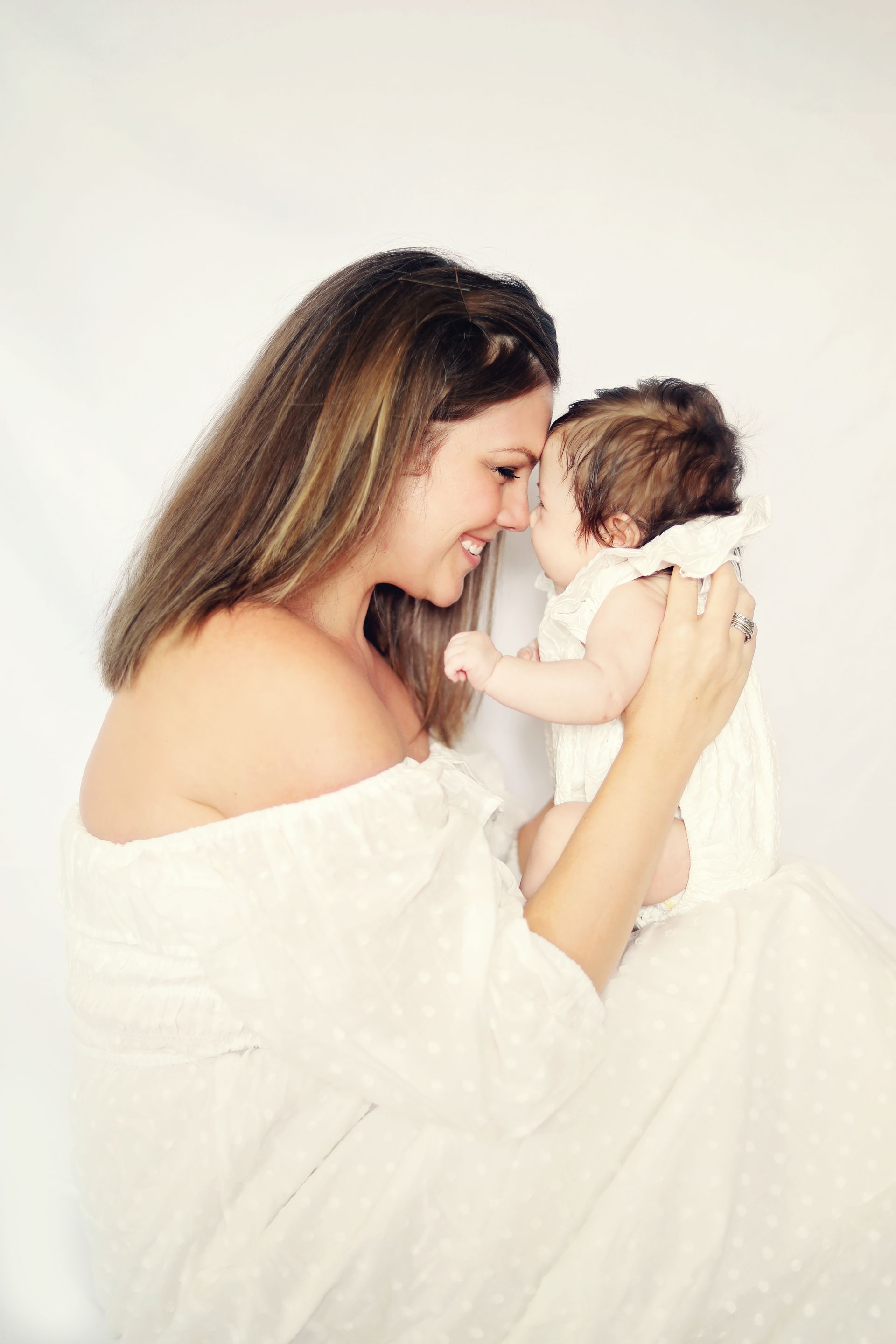 Woman in a white dress gently touching foreheads with a newborn wrapped in white.