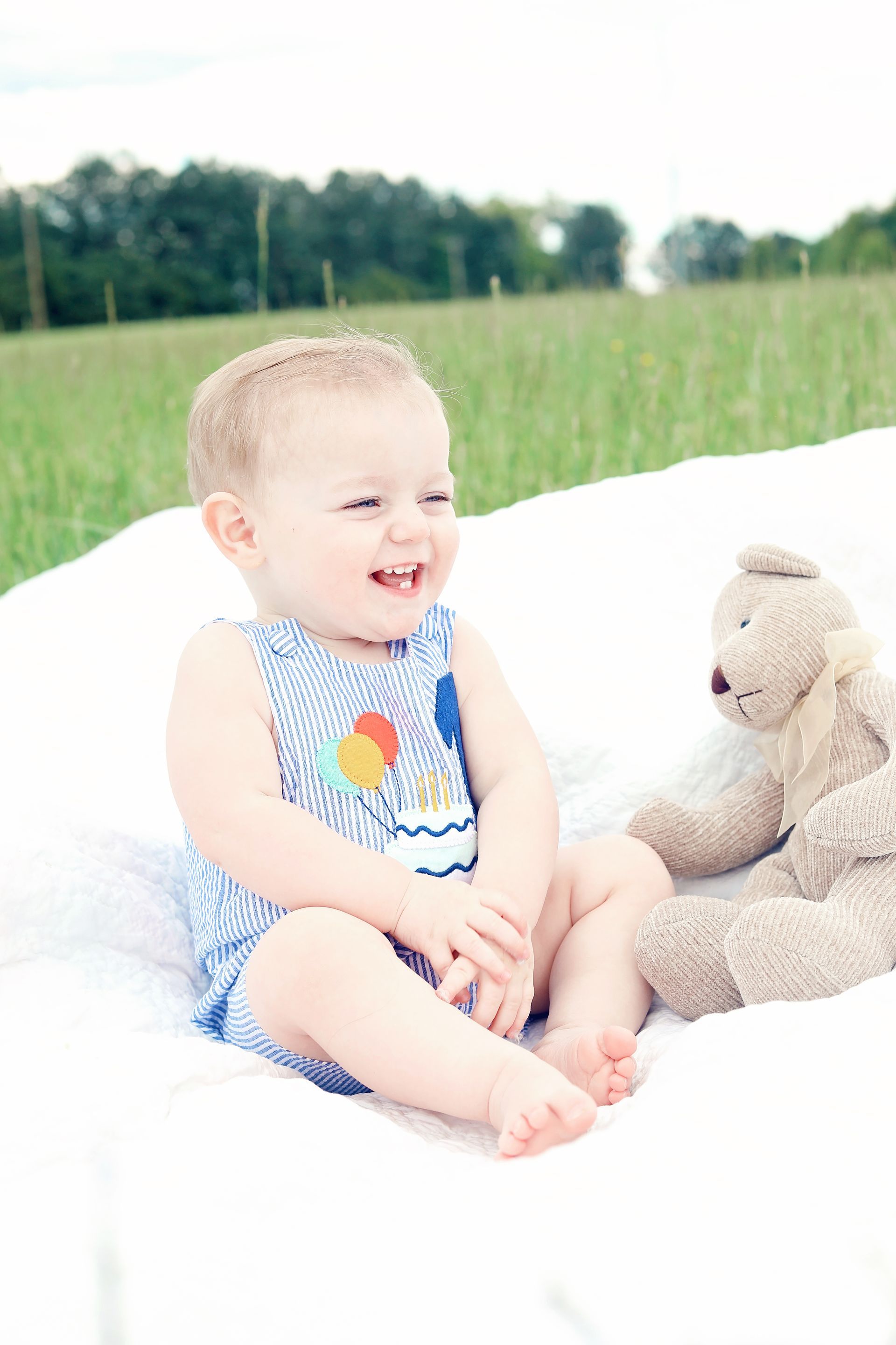 Smiling baby sitting on a white blanket beside a teddy bear in a grassy field