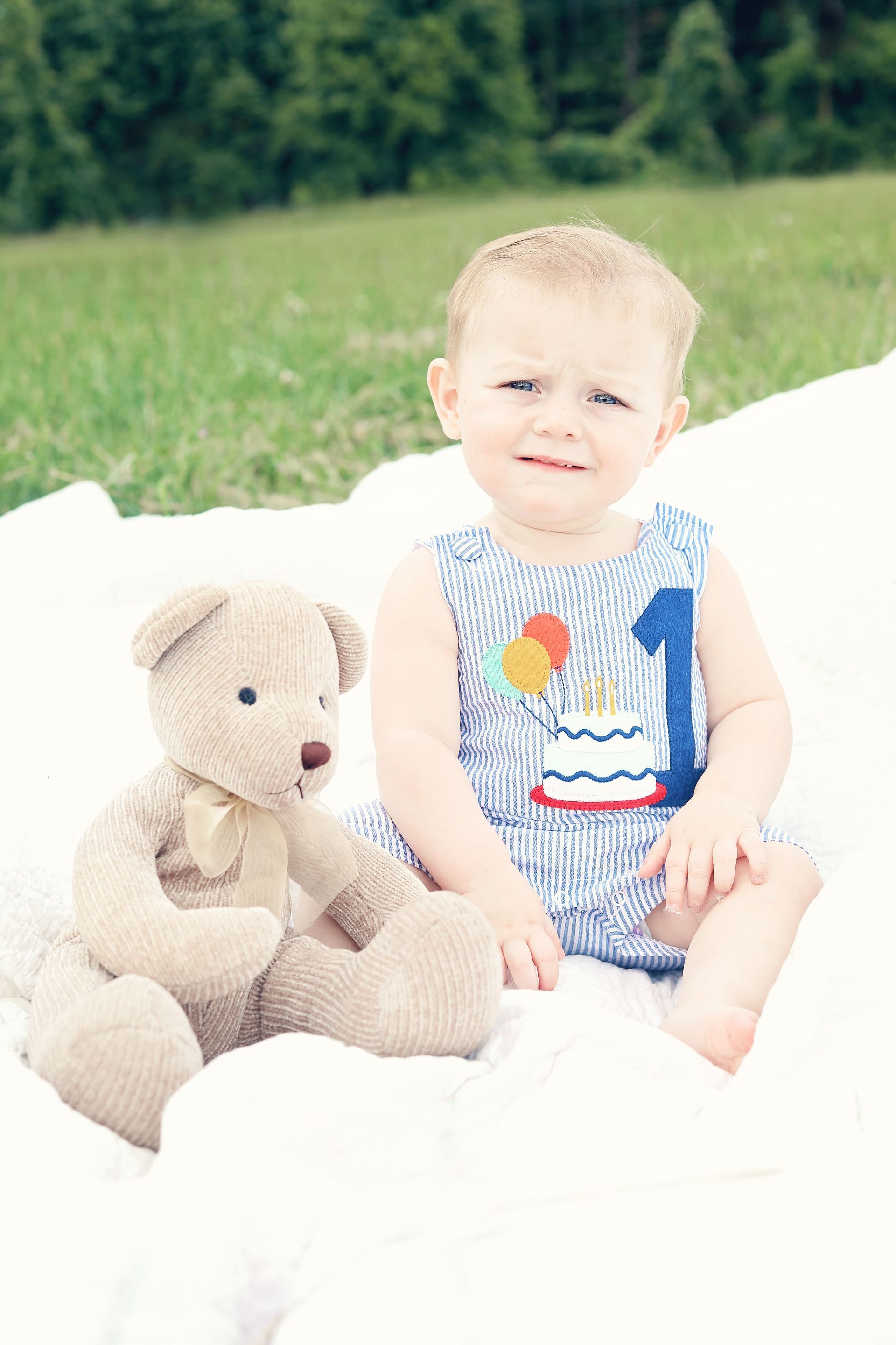 Toddler sitting on a white blanket beside a teddy bear, with green grass in the background.