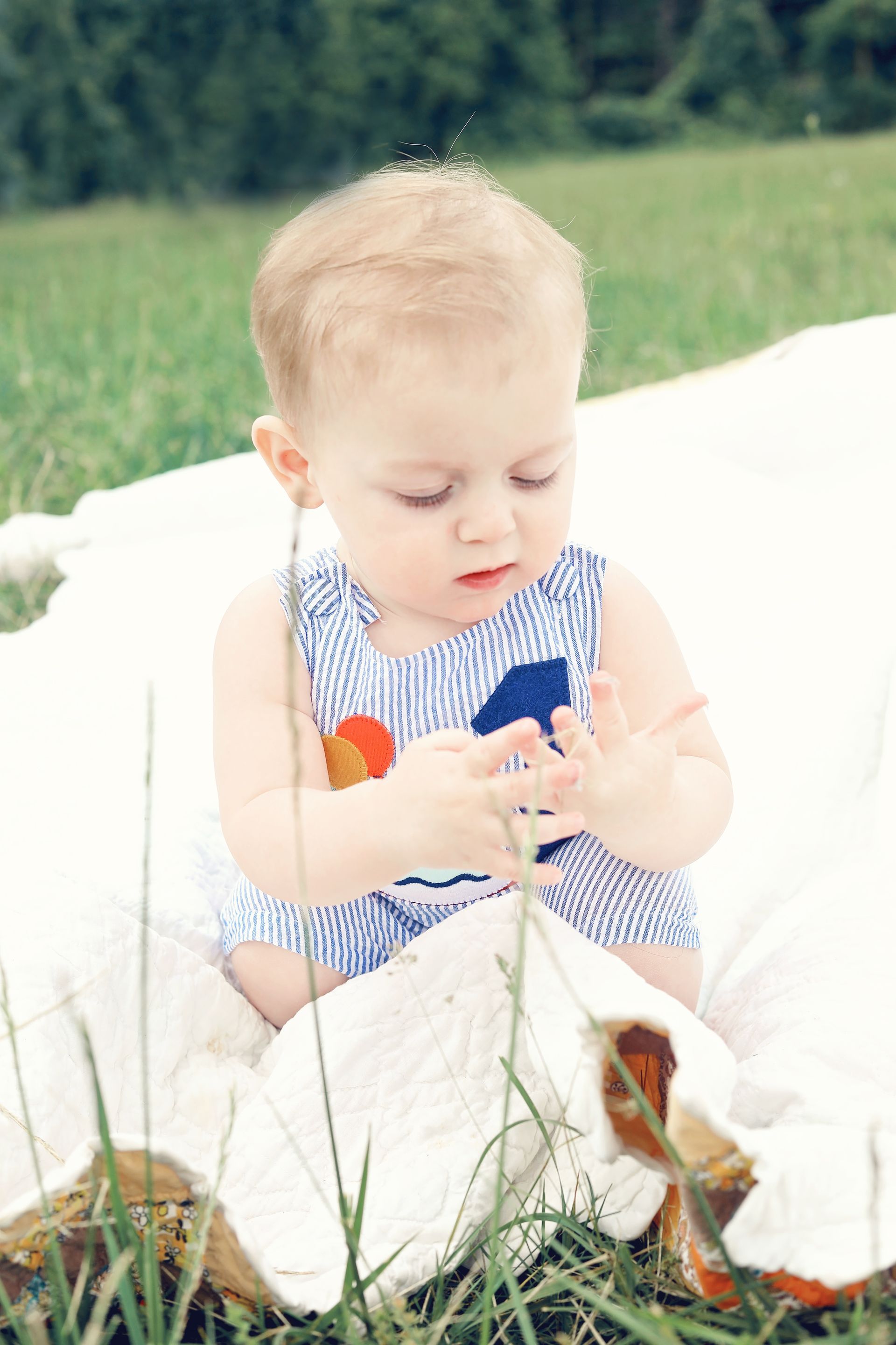 Baby in a blue-and-white outfit sitting on a white blanket, looking at their hands outdoors.