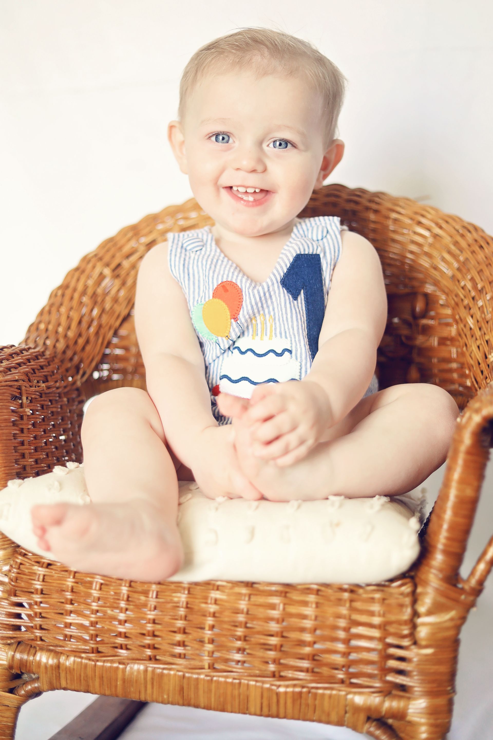 Smiling baby sitting in a wicker chair, wearing a white patterned onesie and resting feet on a cushion.