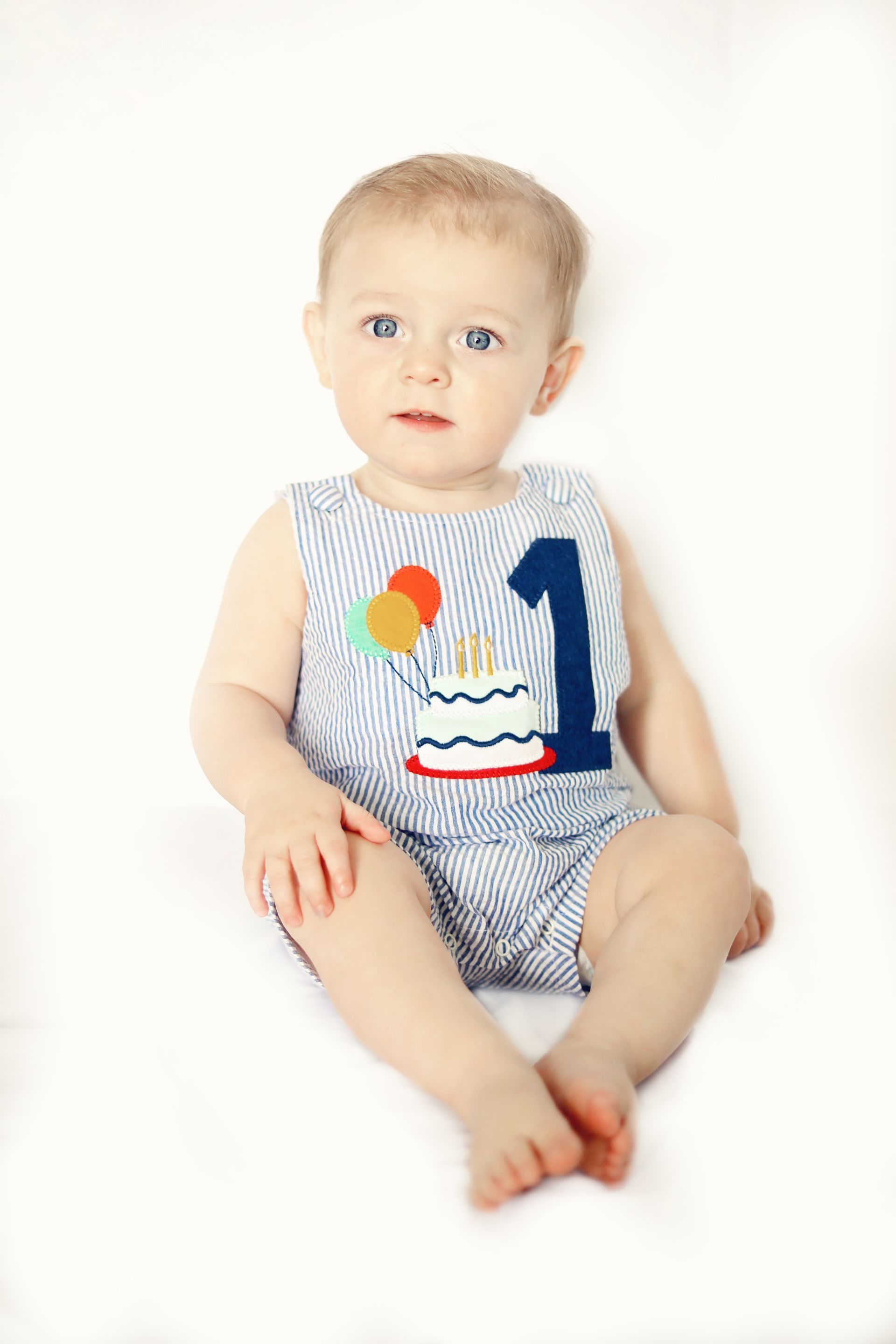 Baby sitting on a white background wearing a patterned romper with a blue number 1 and cake graphic