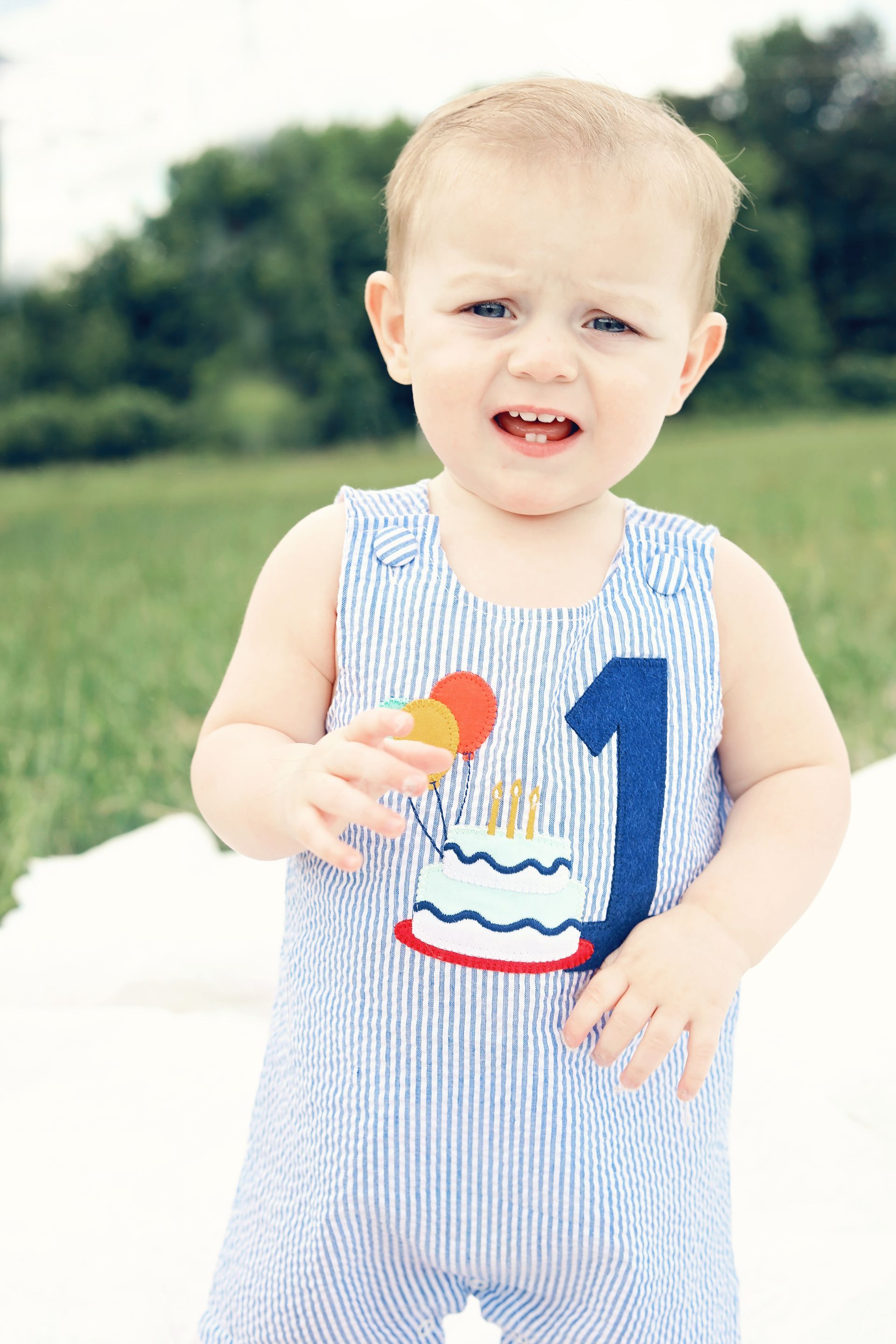 Toddler in a striped romper holding a red-yellow lollipop outdoors, smiling on a sidewalk with green grass behind.
