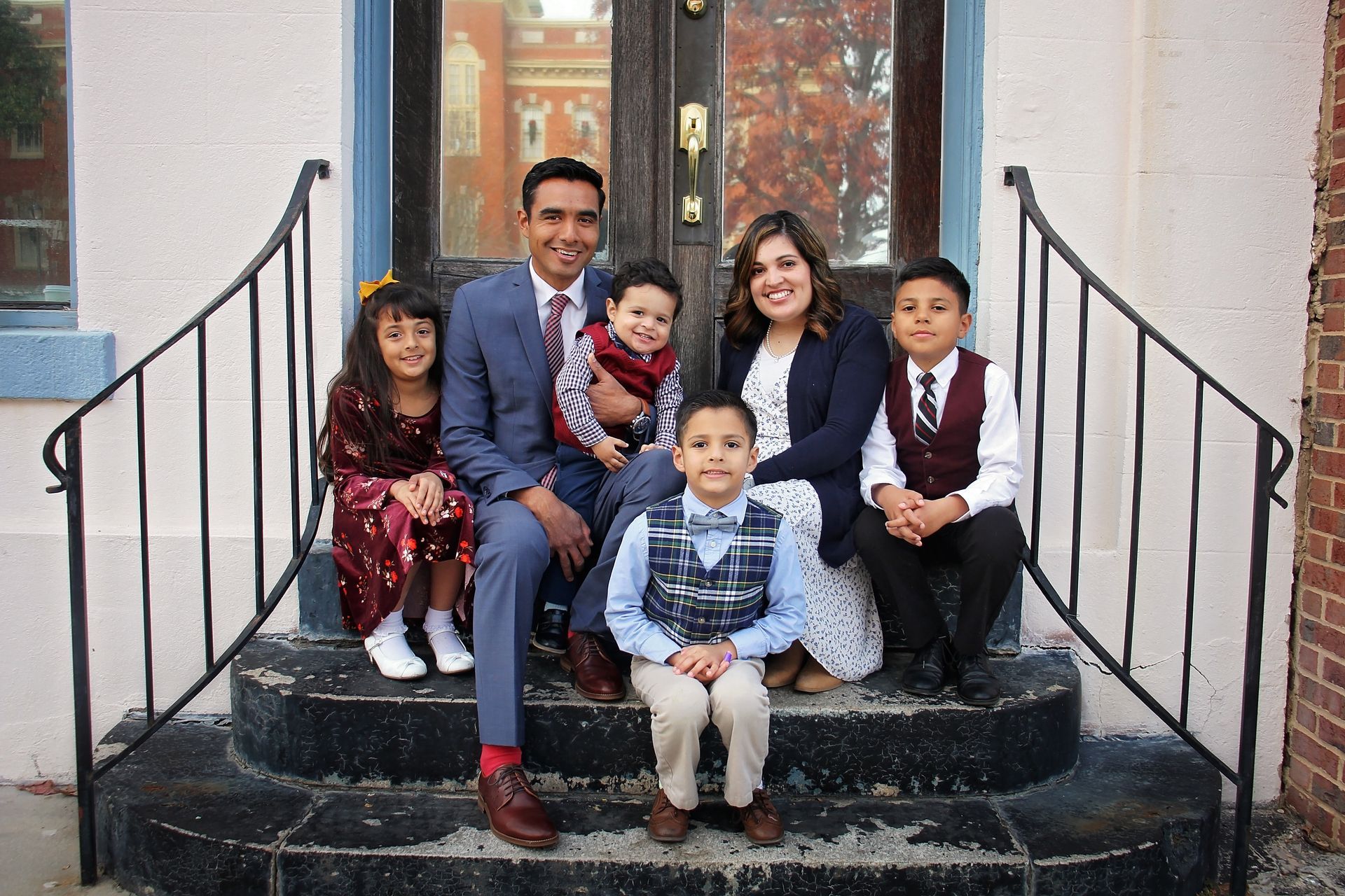Family of five sitting on front steps, smiling in formal clothes, outdoors by a doorway