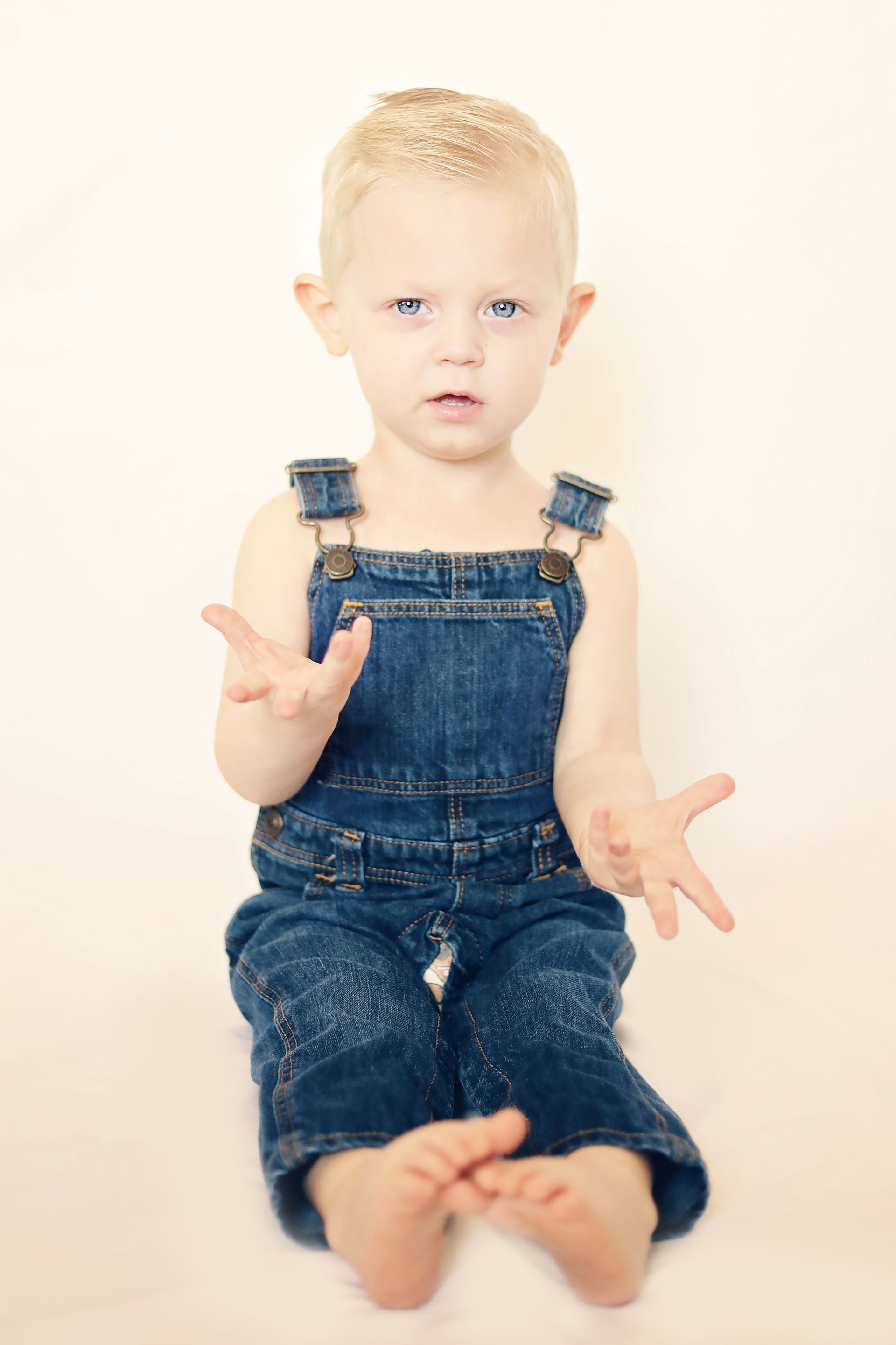Small child in denim overalls sitting on a white background, holding both hands out.