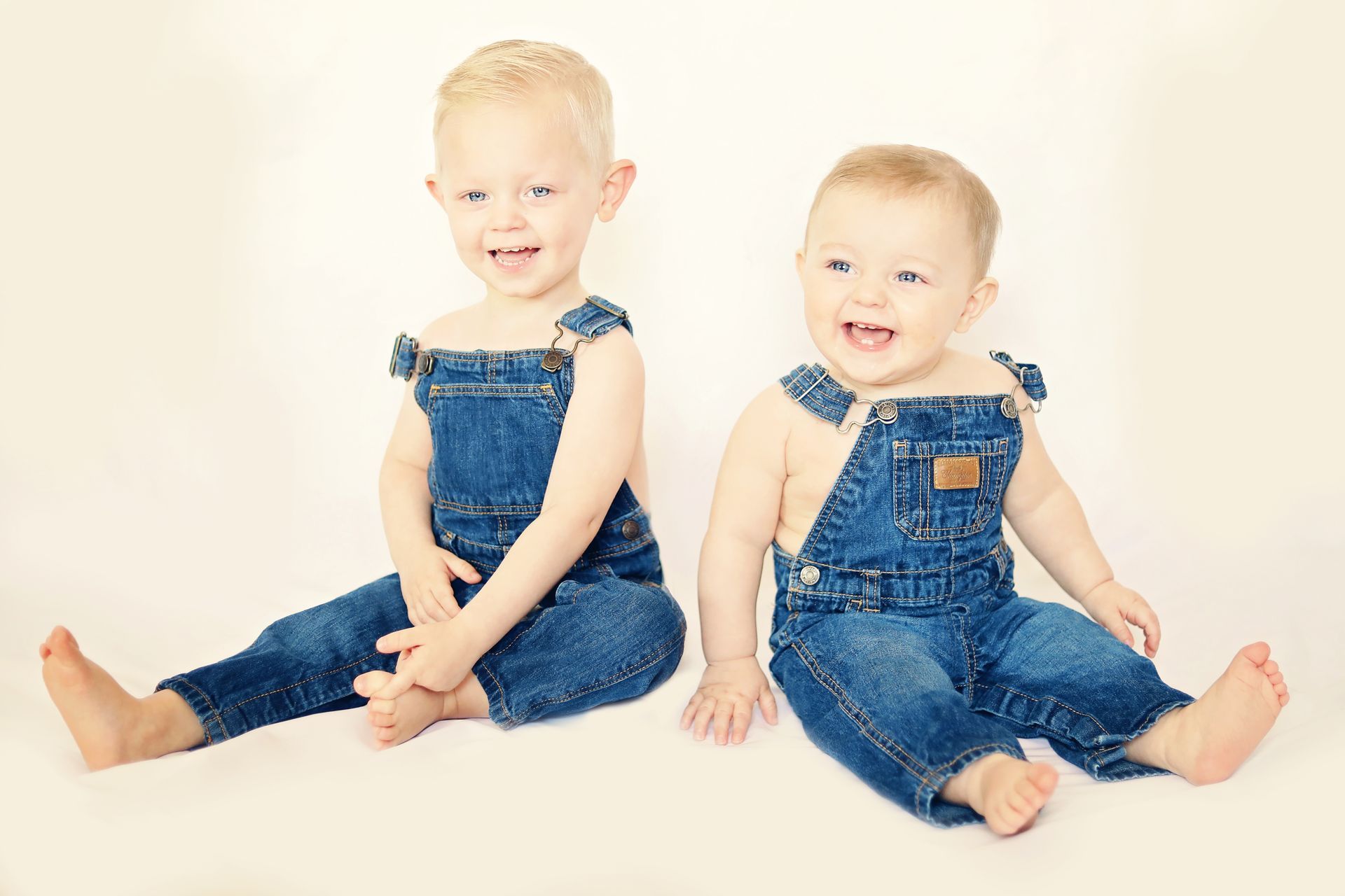 Two smiling toddlers in denim overalls sitting on a white background