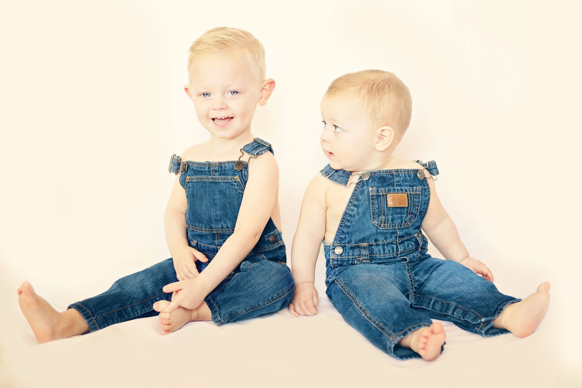 Two toddlers in denim overalls sitting on a light background, one smiling at the camera