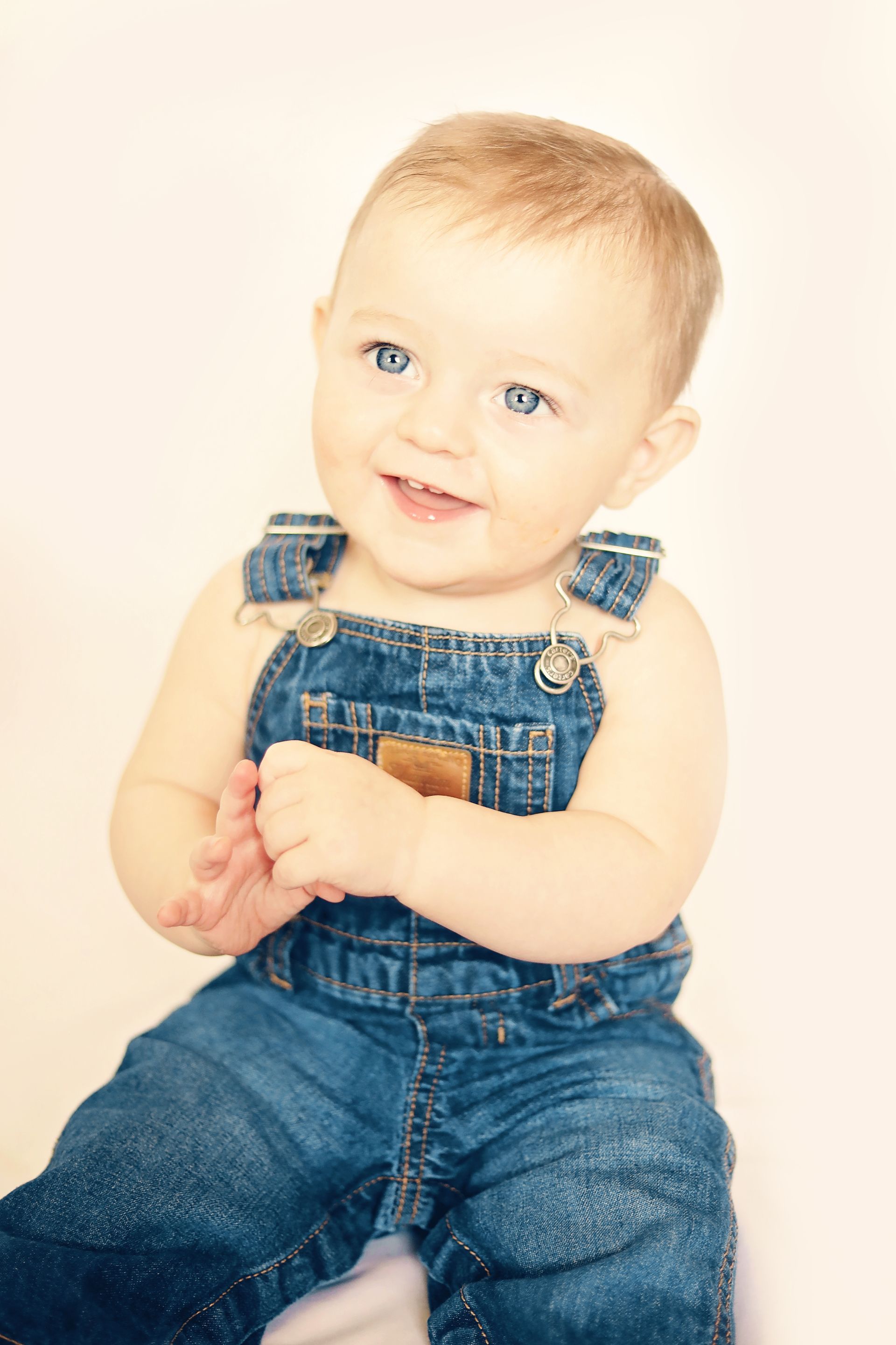 Smiling baby in blue denim overalls, sitting against a light background.