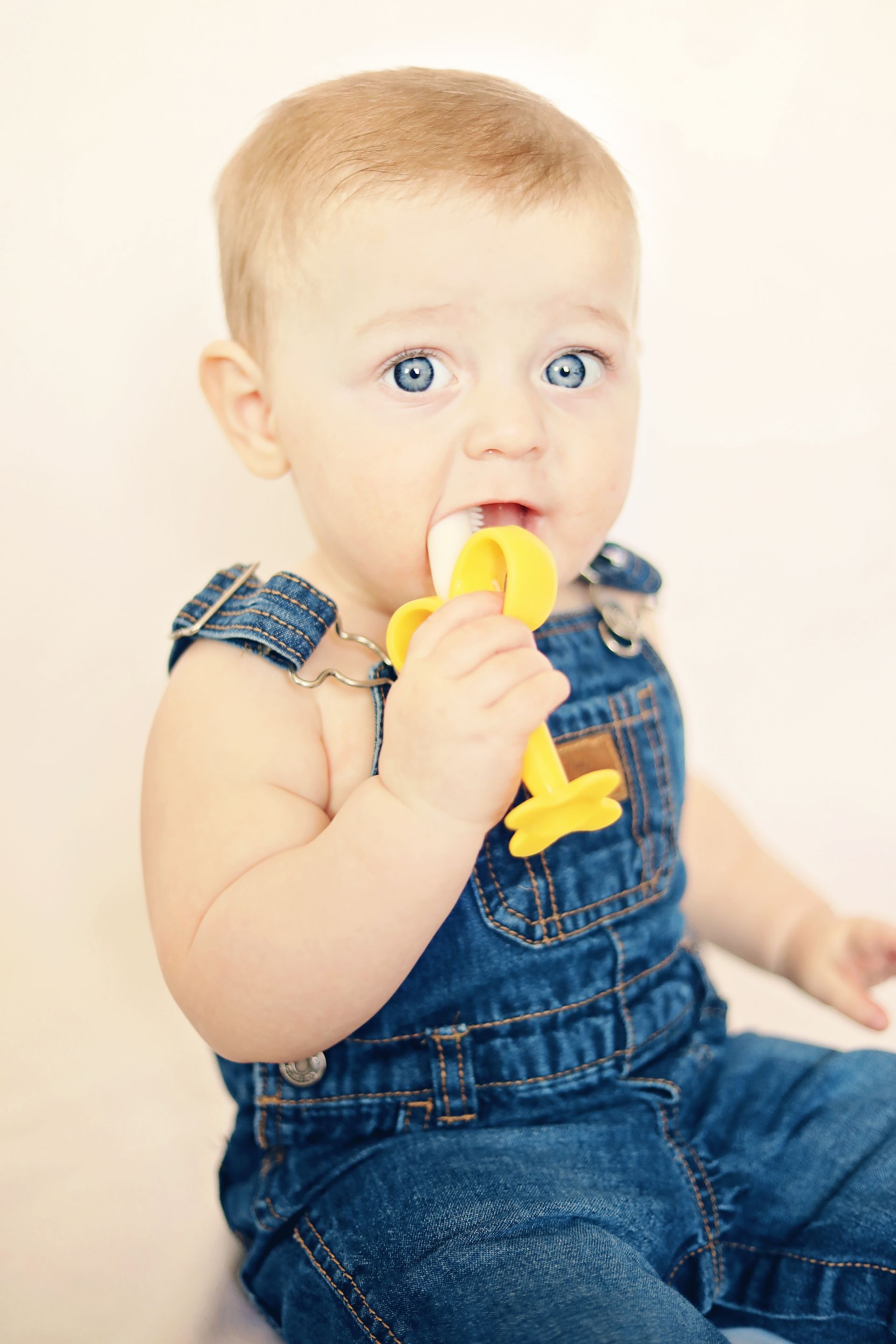 Baby in denim overalls chewing a yellow toy, seated against a soft white background