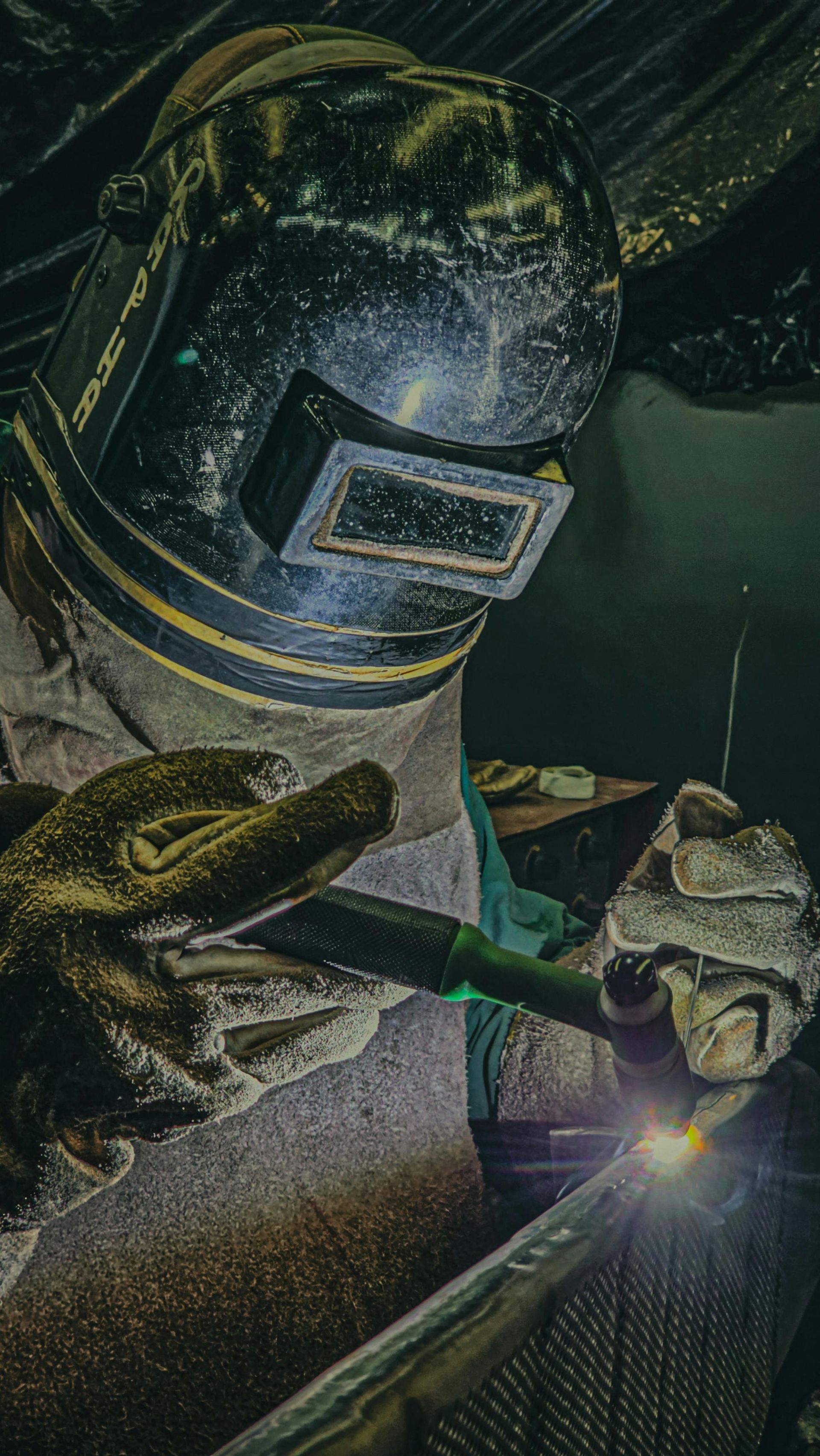 Welder in a dark setting, wearing protective gear and welding metal, sparks visible.