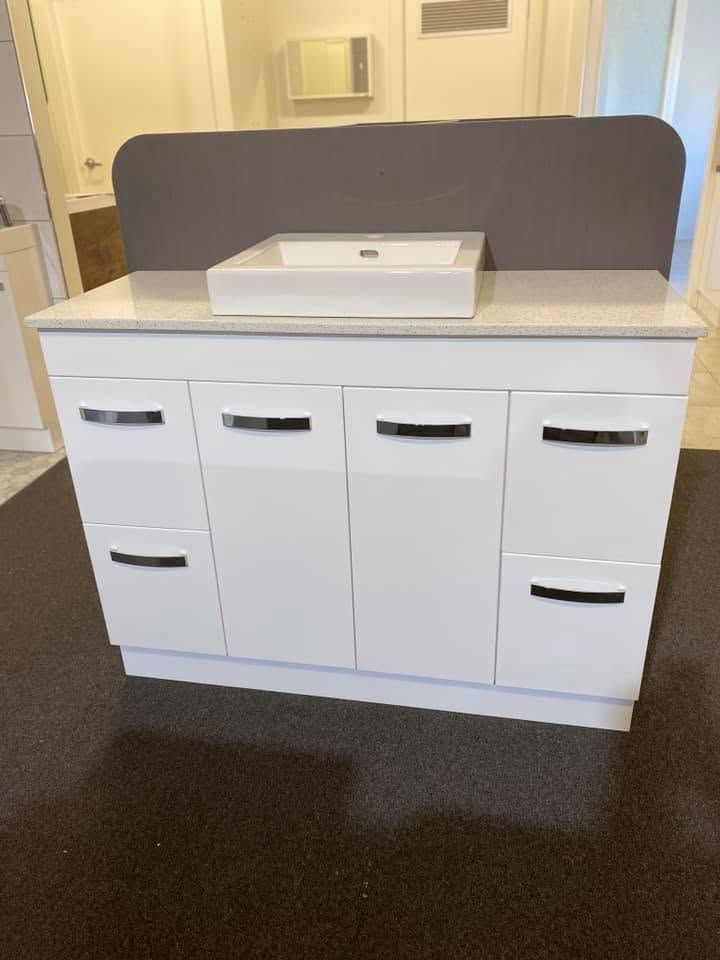 White bathroom vanity cabinet with multiple drawers and a sink, against a dark carpeted floor.