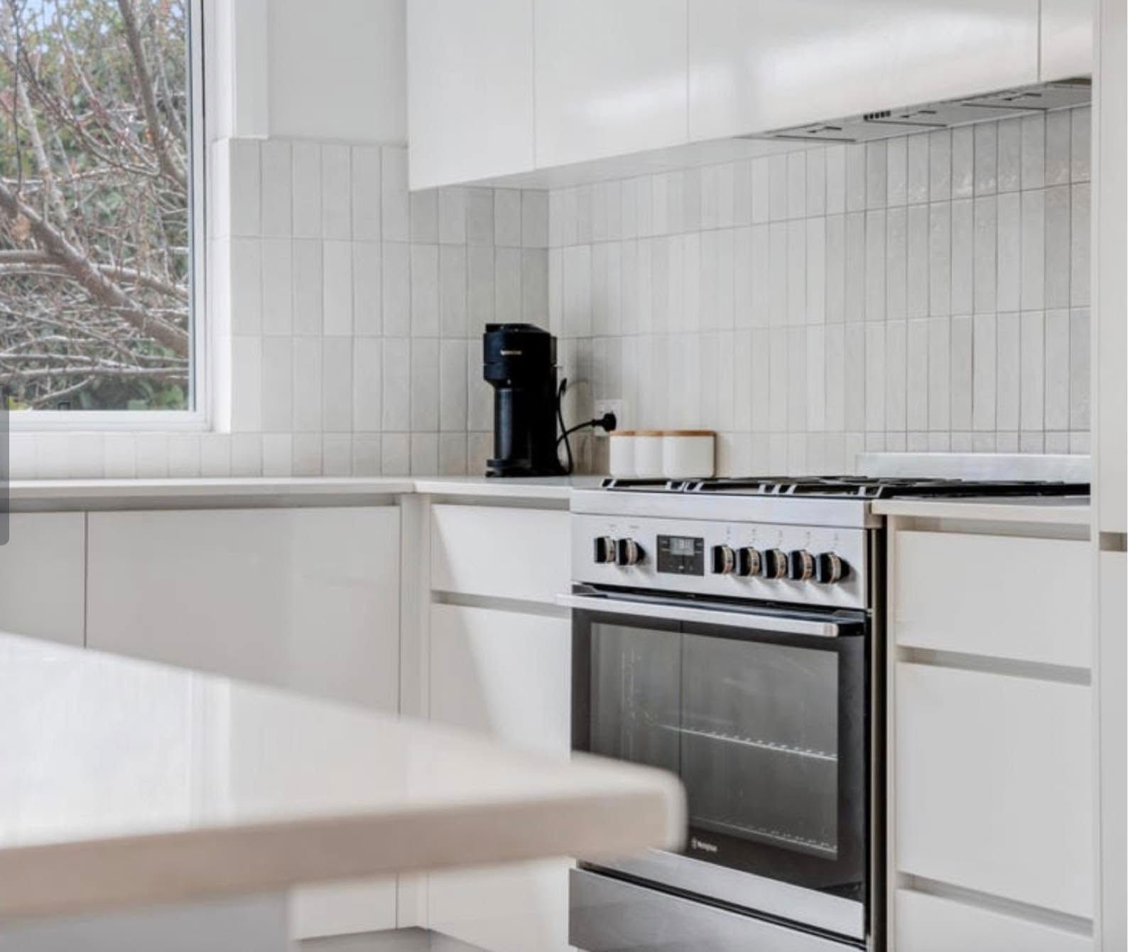 Modern white kitchen with a stove, black coffee maker, and large window beside a white table