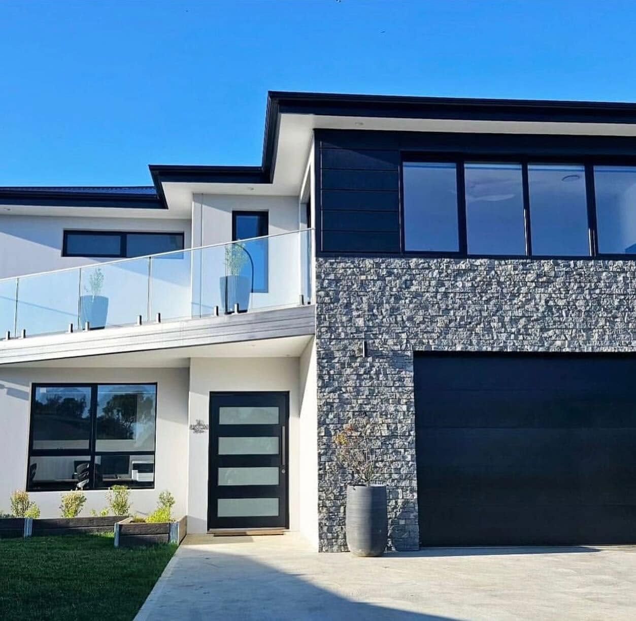 Modern two-story house with white and gray exterior, glass balcony, and large black garage door Modern two-story house with white and gray exterior, glass balcony, and large black garage door