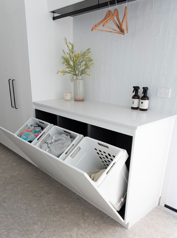 Open white storage cabinet with tilted compartments under a white shelf, with plants, hangers, and bottles above