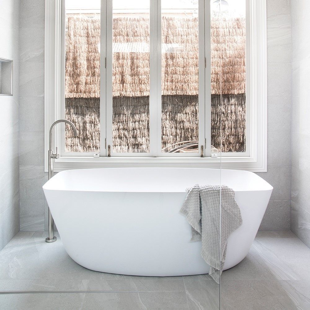 Modern white bathroom with freestanding tub, towel, and large window overlooking a textured wall
