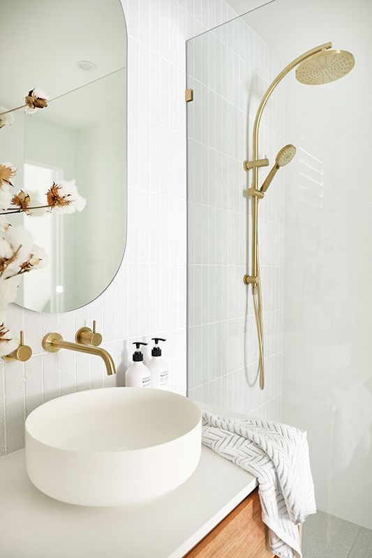 Modern white bathroom with gold fixtures, oval mirror, round sink, and walk-in shower.