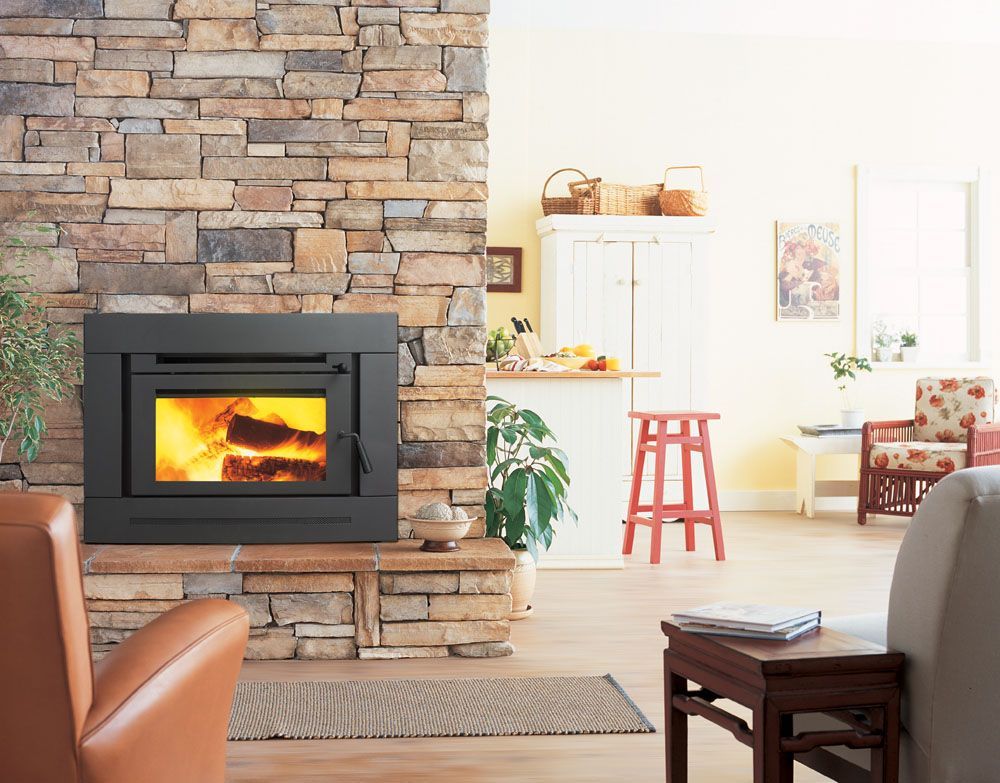 Cozy living room with stone fireplace, lit wood stove, and bright kitchen in the background