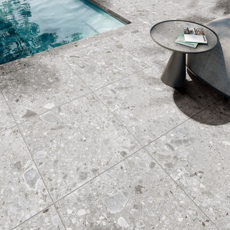 Stone pool deck beside turquoise water, with a small round side table holding books and sunglasses.