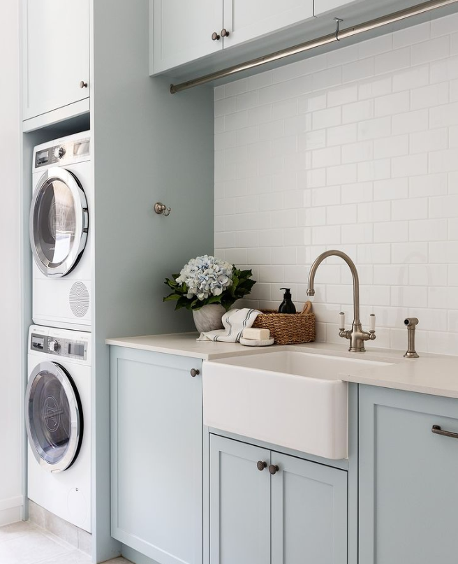 Light blue laundry room with stacked washer-dryer, white sink, brass faucet, and countertop decor