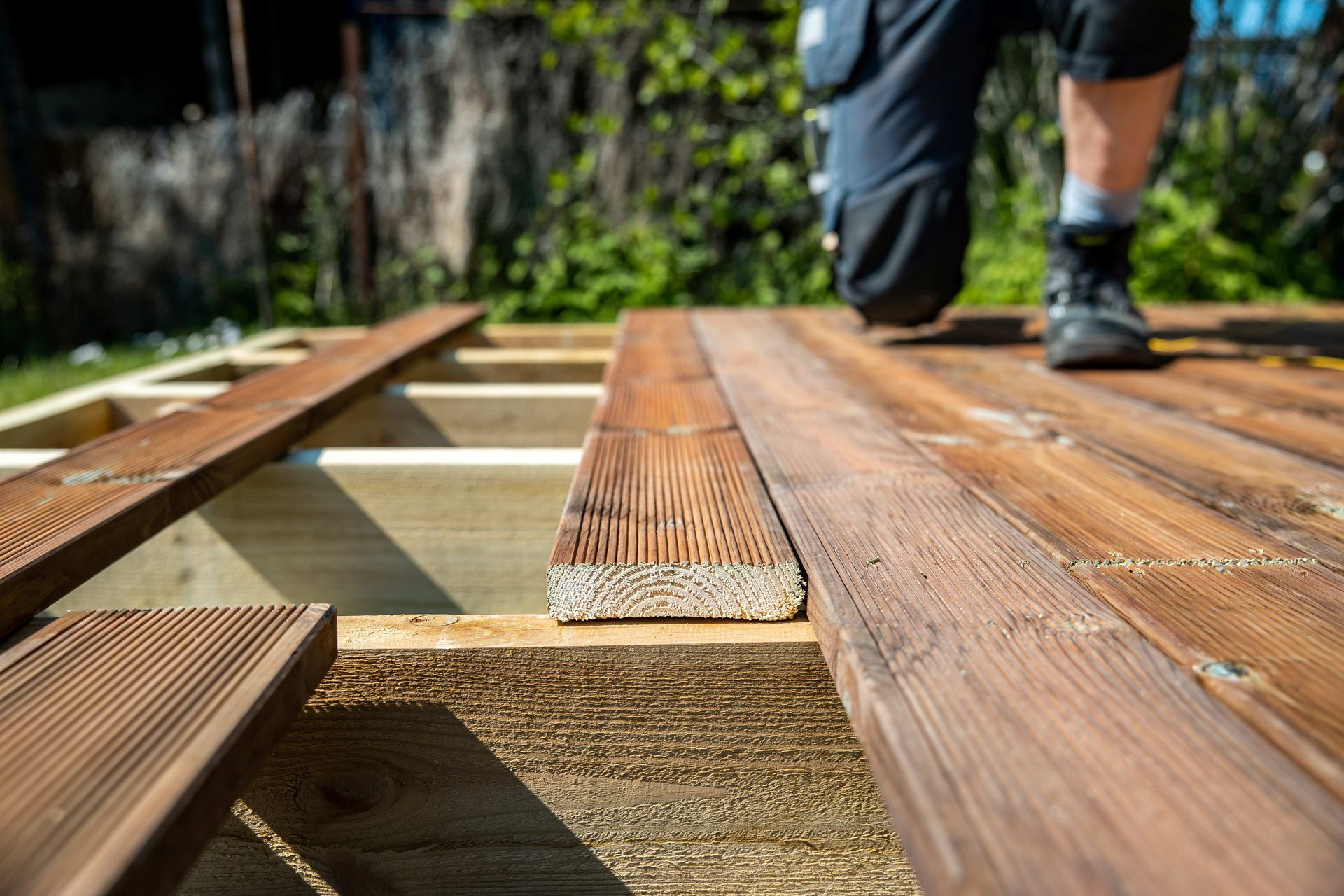 Person installing wooden deck boards outdoors, with tools and framing visible Person installing wooden deck boards outdoors, with tools and framing visible