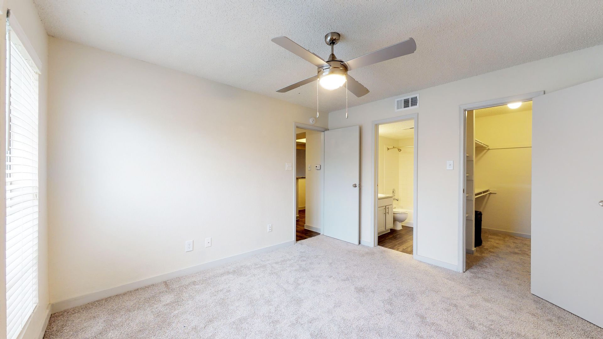 Empty beige bedroom with carpet, ceiling fan, and two open doorways to a bathroom and closet.