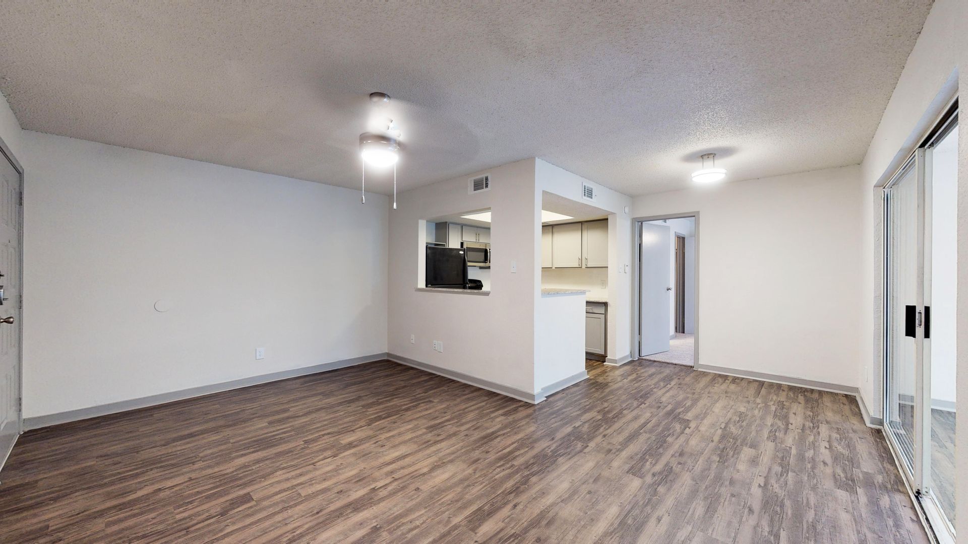 Empty living room with wood floors, white walls, and sliding glass doors, leading to a small kitchen nook