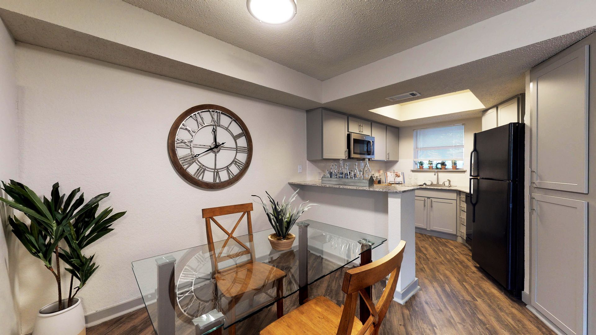 Modern dining area with glass table, wall clock, kitchen, and stainless steel refrigerator