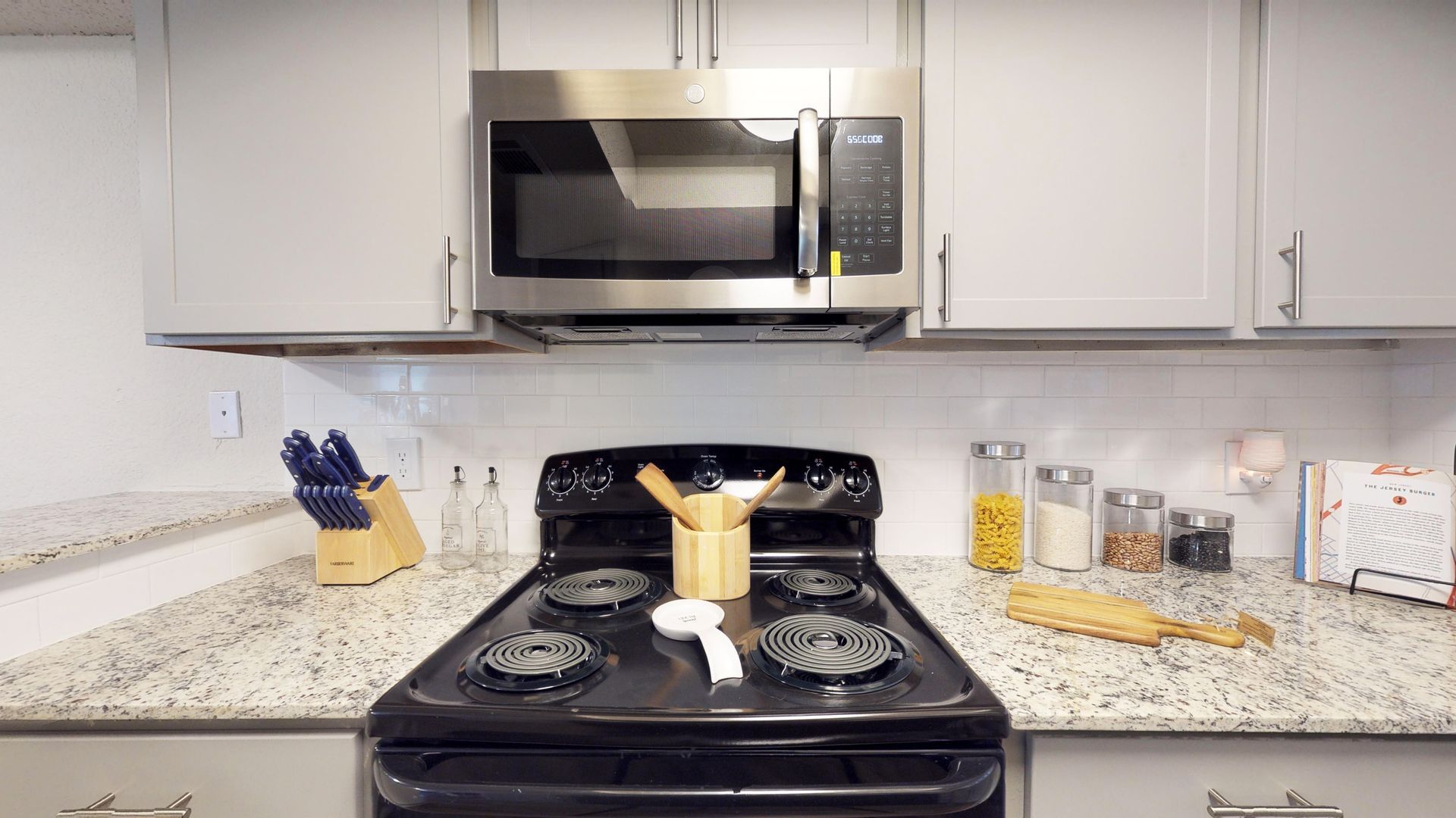 Kitchen with black stove and microwave, white cabinets, speckled counters, and utensil holders on either side