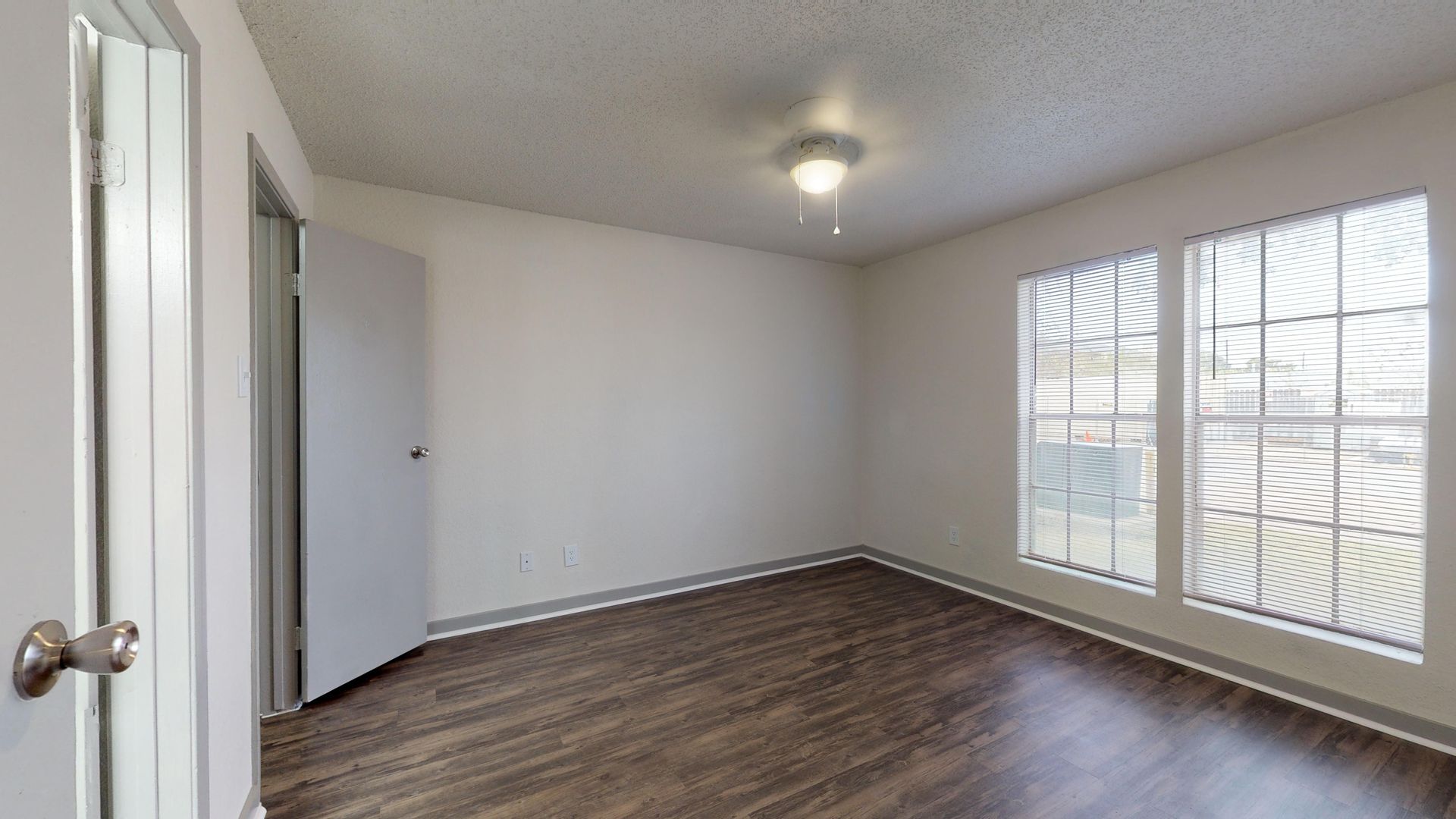 Empty bedroom with wood floors, white walls, ceiling light, and two windows with blinds