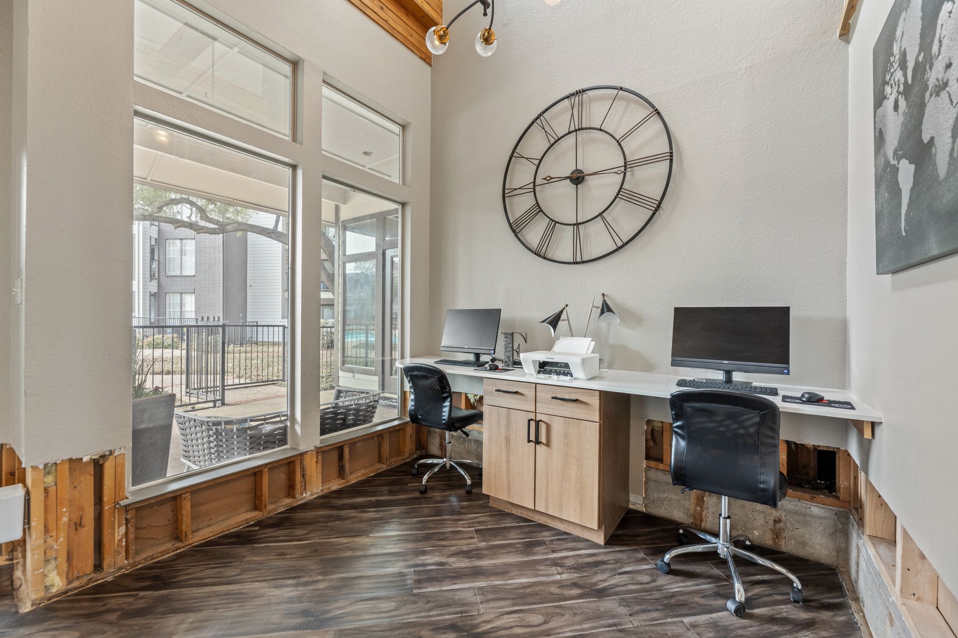 Modern office corner with two desks, computers, black chairs, large wall clock, and tall windows