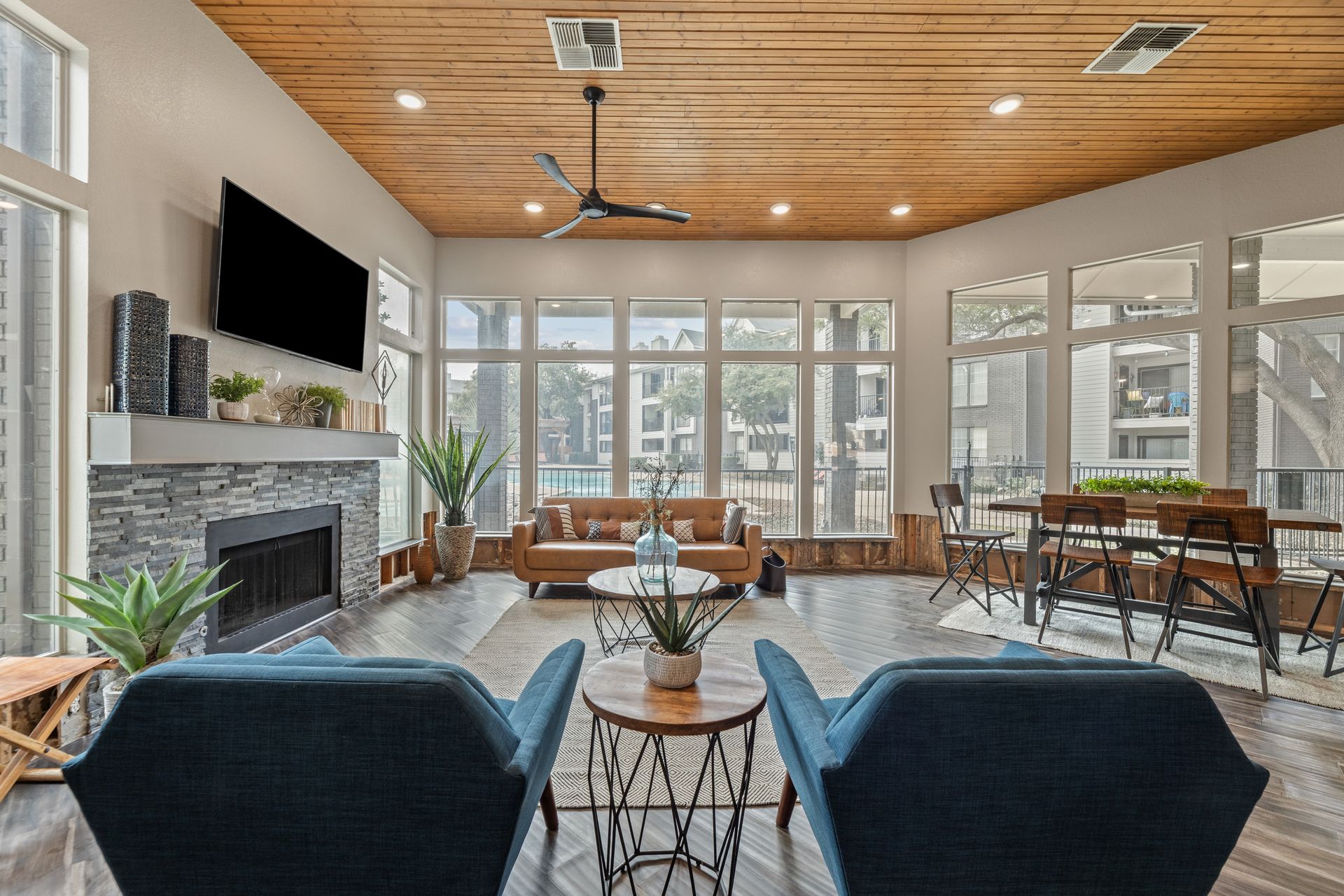Bright modern sunroom with blue chairs, fireplace, large windows, wood ceiling, and patio dining area