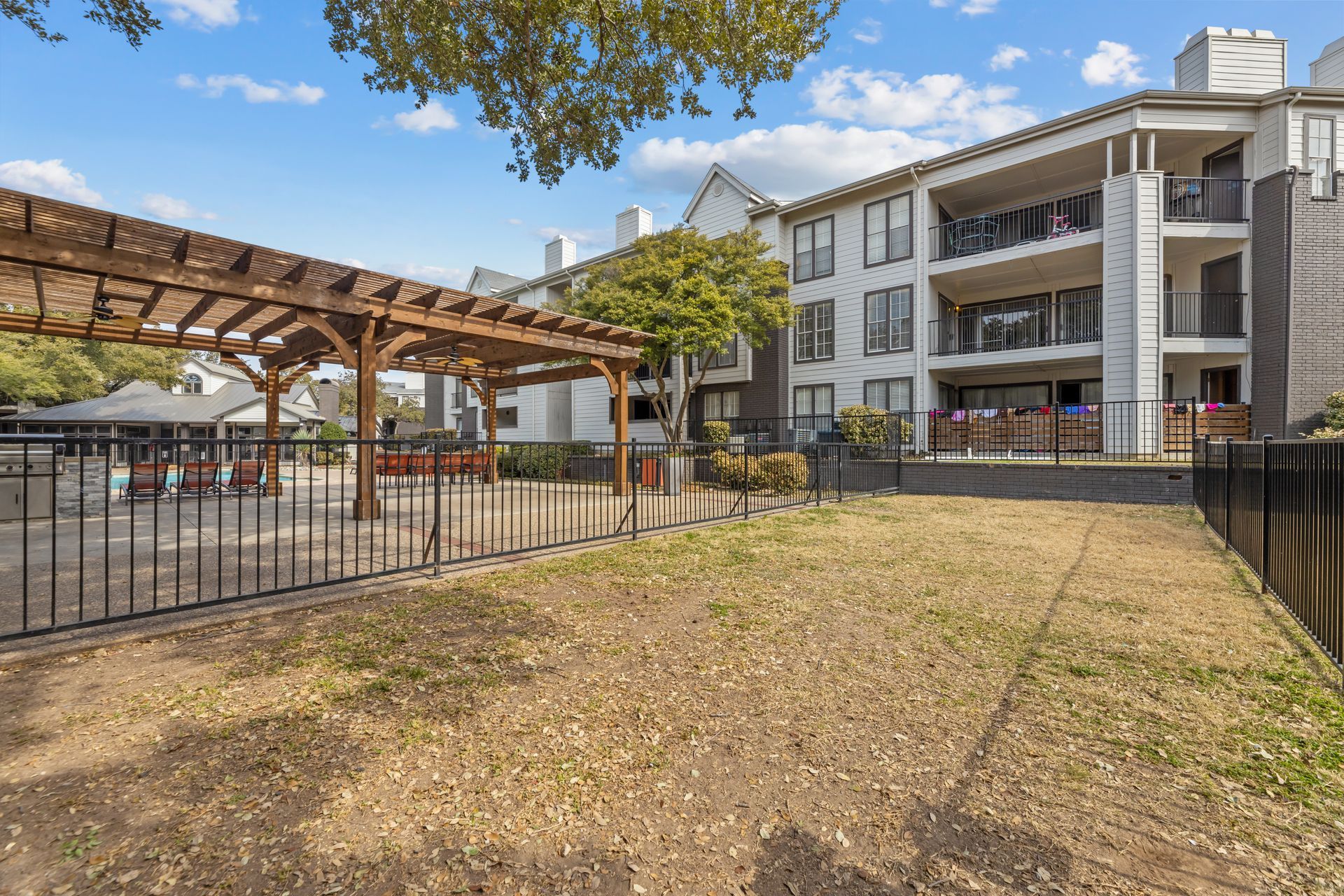 Apartment courtyard with dry grass, wooden pergola, and a multi-story building under a blue sky