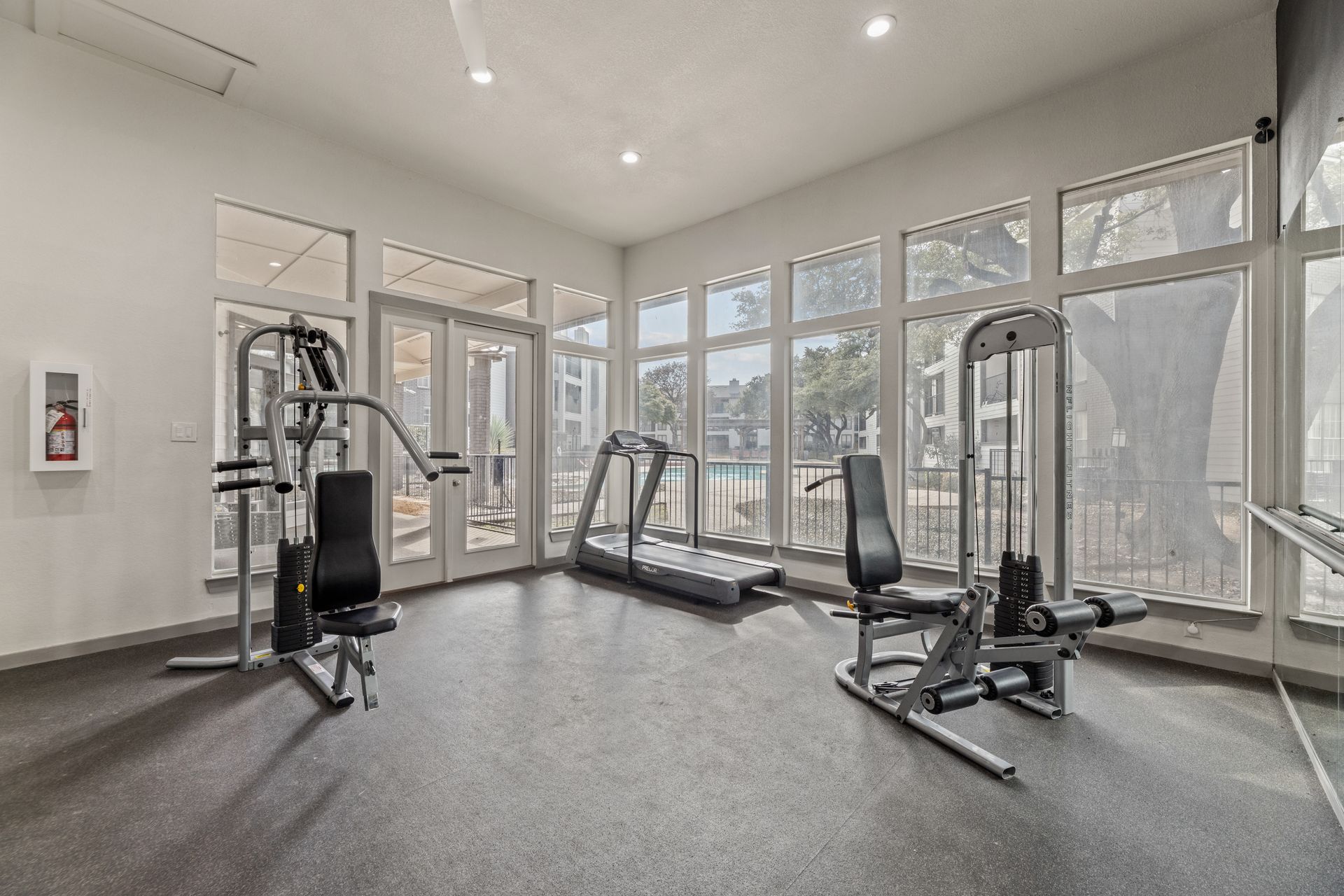 Bright gym room with cardio and strength machines, gray carpet, and large windows.