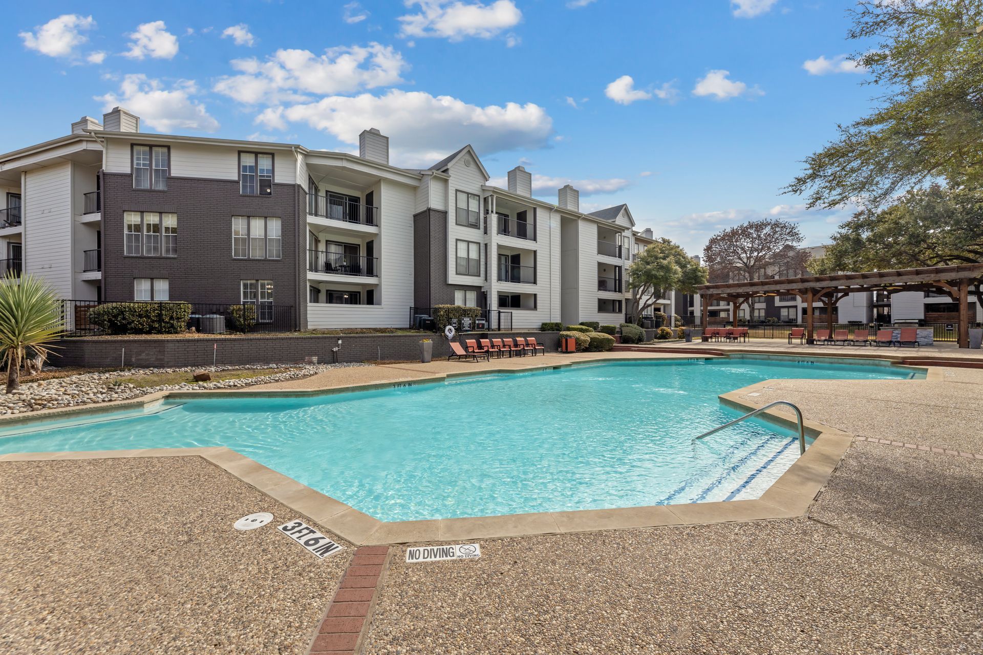 Outdoor apartment complex pool with turquoise water, lounge chairs, and blue sky with clouds