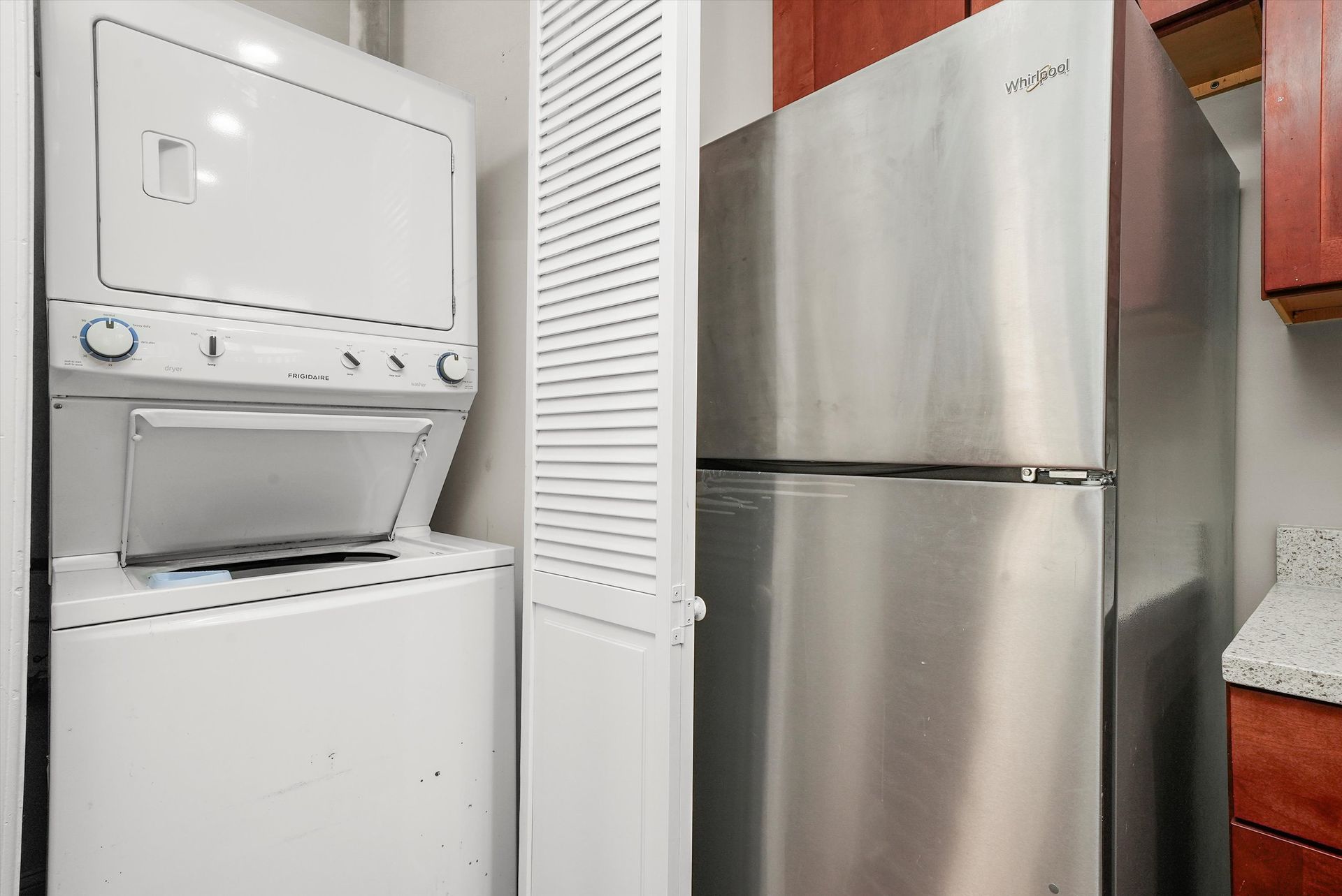 Stackable white washer/dryer, tall stainless steel refrigerator, and a louvered door in a small kitchen.