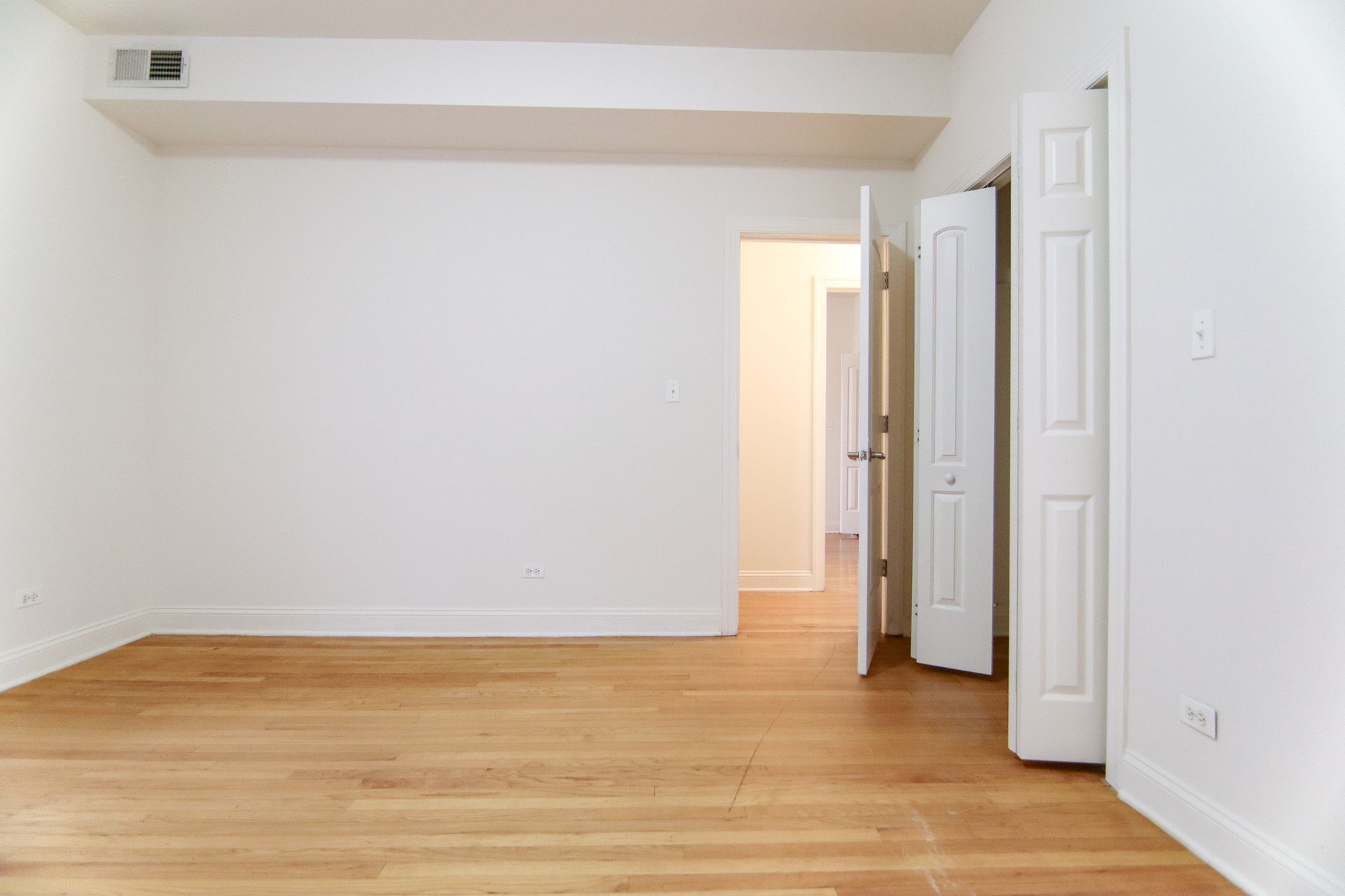 Empty room with hardwood floors and white walls. Two doors open to hallway and closet.
