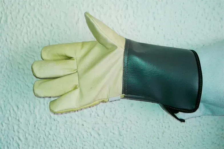 Una mano con un guante de cuero beige y un puño largo de color gris oscuro sobre un fondo blanco texturizado.