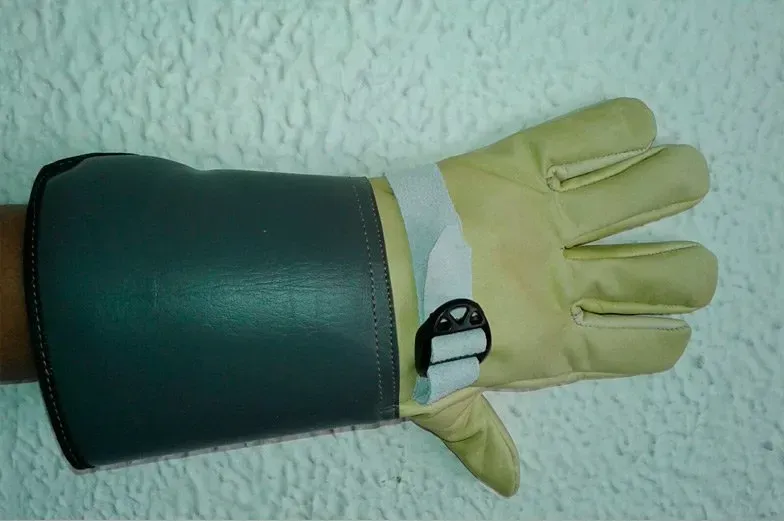 Un guante reforzado de color marrón claro con un puño protector verde oscuro y una correa blanca ajustable, que se lleva en la mano.