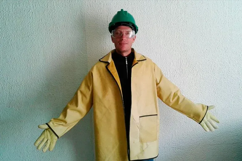 Una persona que lleva un casco verde, gafas de seguridad, una chaqueta protectora amarilla y guantes amarillos está de pie apoyada contra una pared.