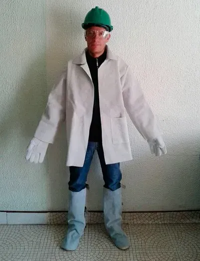 Una persona con casco verde, gafas protectoras, bata blanca, guantes de trabajo y cubrepiernas grises, apoyada contra una pared blanca.