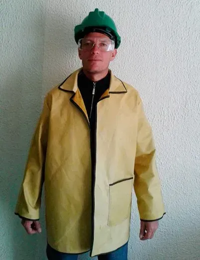 Una persona con casco verde, gafas protectoras y chaqueta de trabajo amarilla de manga larga, apoyada contra una pared lisa.