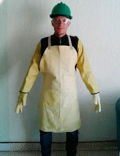 Una persona que lleva un casco verde, gafas de seguridad, mangas protectoras amarillas, guantes y un delantal químico beige.