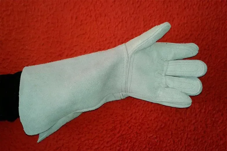 Un guante de soldar de cuero blanco, tipo manopla, que se lleva puesto en una mano sobre un fondo rojo.