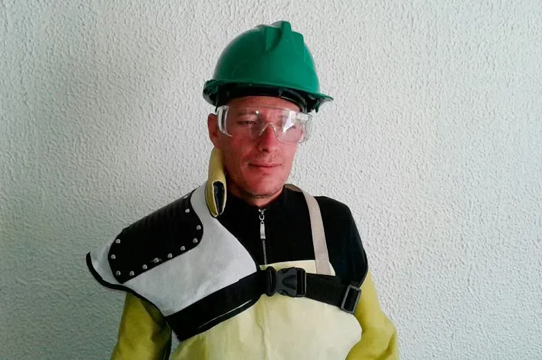 Una persona lleva un casco verde, gafas de seguridad y un traje protector de cuero sobre una camisa negra, apoyada contra una pared blanca.