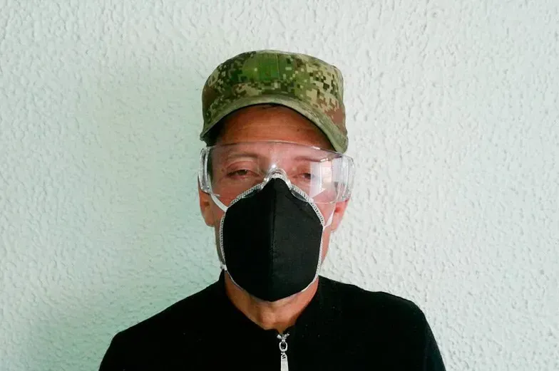 Una persona con gorra de camuflaje, gafas de seguridad transparentes y mascarilla negra frente a una pared texturizada de color verde pálido.