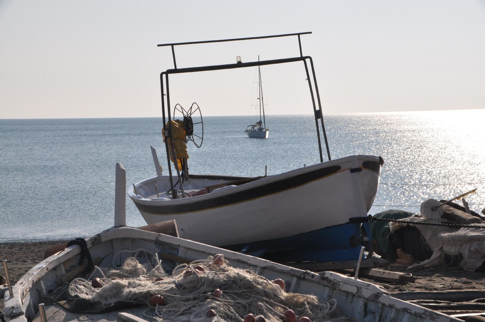 Barca da pesca bianca su una spiaggia, struttura metallica che si inarca sopra la testa, barca in lontananza, luce del sole splendente sull'acqua.